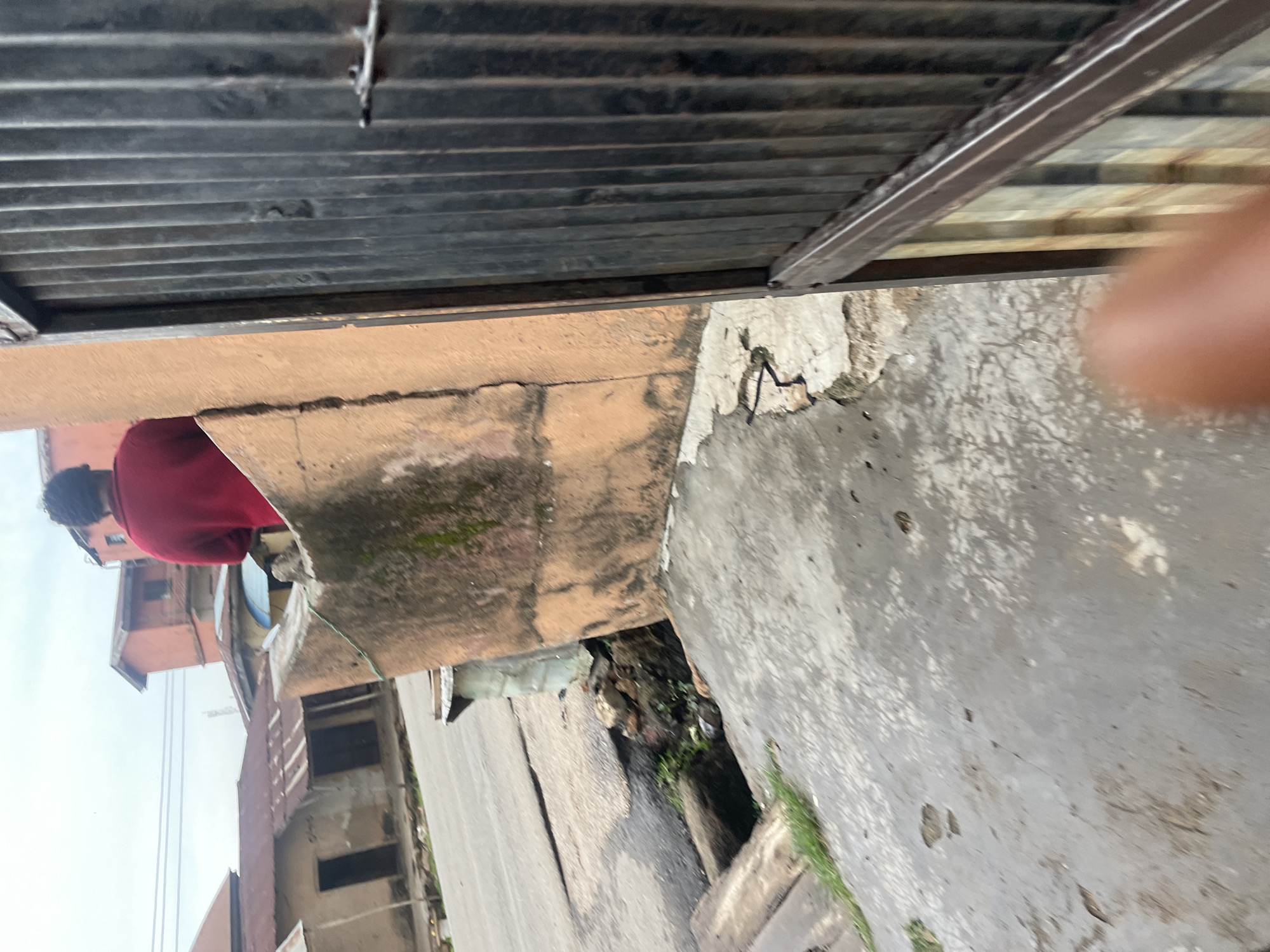 The image shows a street scene with damaged infrastructure in Jos, Nigeria. In the foreground, a damaged concrete surface is visible, with a section of the road appearing to have collapsed into a ditch. Debris and vegetation are present in the ditch. A person wearing a red shirt is standing on a dilapidated wall in the background, facing away from the camera. Behind the wall, several buildings with corrugated iron roofs are visible, typical of residential areas in Jos. A metal gate or fence is partially visible on the right side of the image. The sky is overcast.