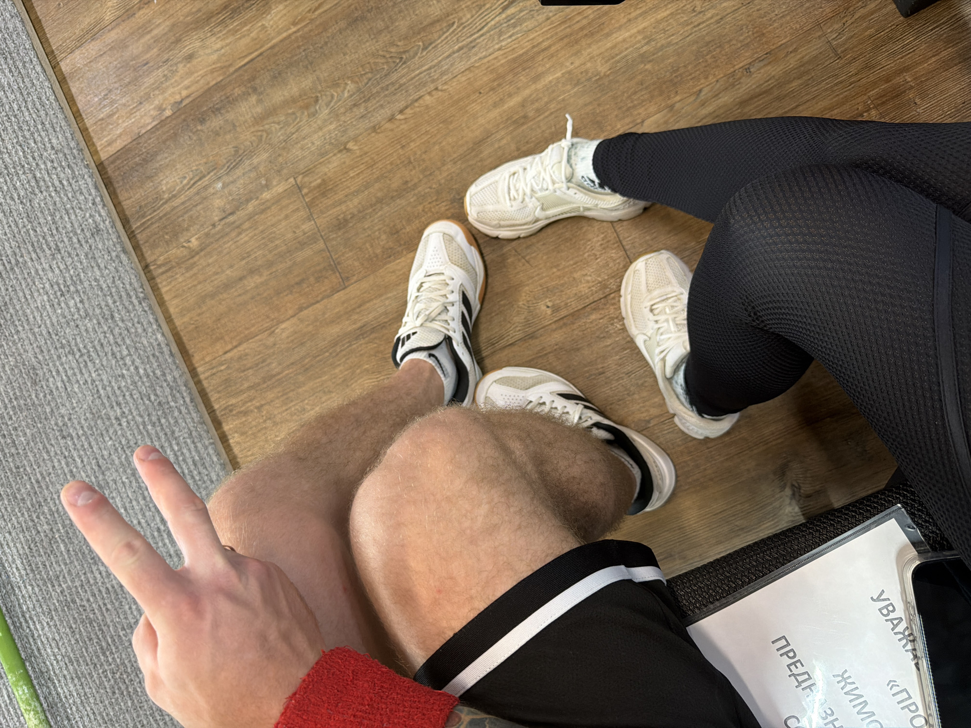 Here is a description of the image:
**People:** Two people are visible in the image. One person is wearing black shorts with white stripes, a red sweater, and tennis shoes. This person is also making a "peace" sign with their hand. The second person is wearing black leggings and white running shoes.
**Objects:** Other objects in the scene include a gray rug, a wood floor, a document in a plastic sleeve, and a black cell phone.
**Actions:** The two people are sitting. One person is making a "peace" sign.
**Environment:** The scene appears to be indoors, possibly in a gym or waiting area. The presence of the document and phone suggests that the people may be waiting or engaged in some kind of administrative task.
**Location:** While the image's origin is mentioned as Chaikovsky, Russia, I lack the ability to confirm the exact location within the city based solely on this image.