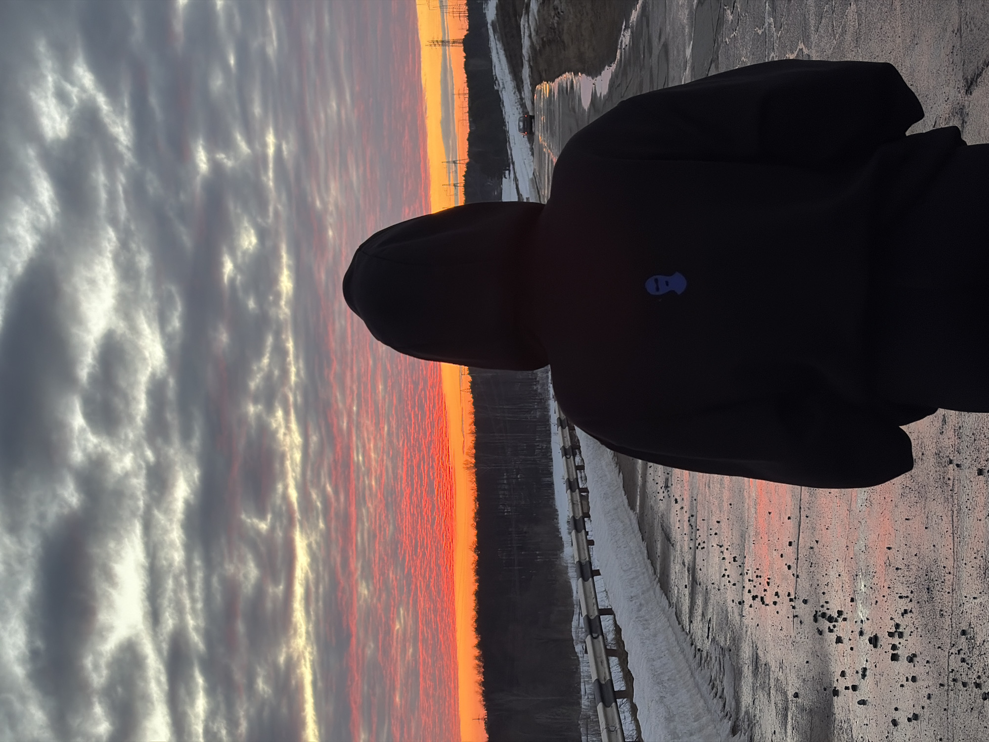 The image displays an individual, viewed from behind, standing on a partially wet paved surface at sunset in Chaikovsky, Russia. The person wears a black hooded sweatshirt with the hood up, revealing a small blue balaclava emblem on the upper back. The sky exhibits a dramatic sunset with intense red and orange hues near the horizon, transitioning to gray and white altocumulus clouds overhead. The wet pavement reflects the sky's colors, and numerous small, dark debris fragments are scattered across it. To the left of the road, a snowbank is present, bordered by a black and silver metal guardrail. Beyond the guardrail, a dense line of trees forms a dark silhouette against the horizon. To the right, the ground slopes upward, featuring patches of snow and bare earth. In the distance on the right, multiple power line transmission towers are visible against the illuminated sky. A dark-colored car is positioned further down the road, away from the viewer.
