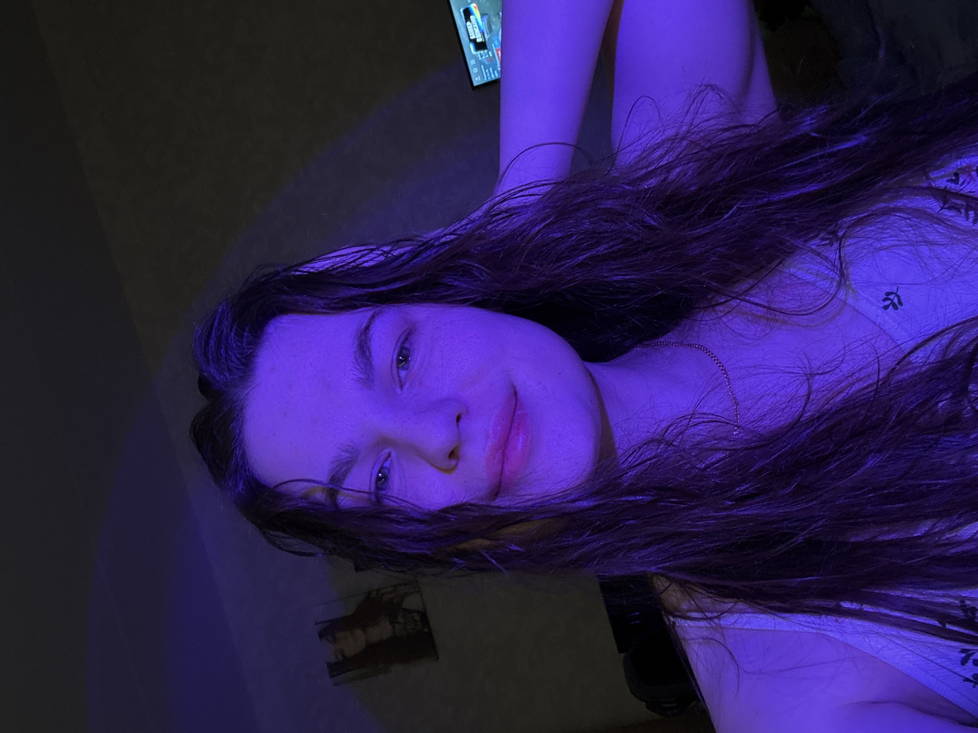 A selfie shows a young woman with long, dark hair and fair skin. She is smiling slightly. She is wearing a white top with a faint floral pattern. The image has a strong blue hue due to the lighting. In the background, there's a wall, a TV screen showing a video game, and a framed picture. The location is unknown. The city of Chaikovsky, Russia is not visible in the image.