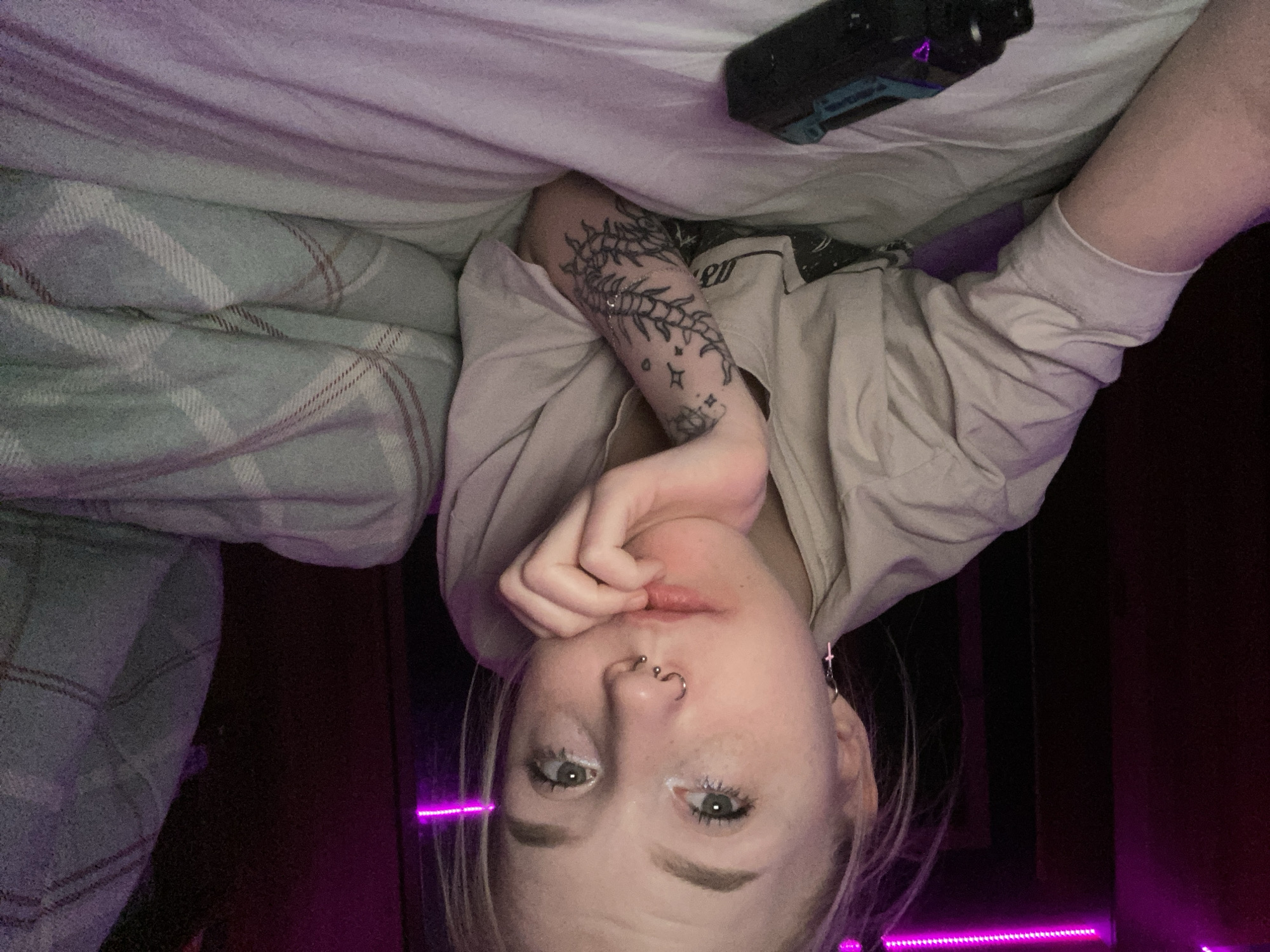 The image depicts a young adult female, positioned in a reclining posture on a bed surface, gazing directly into the camera. She possesses light-colored hair, a horseshoe barbell piercing in her septum, a stud or ring in her right nostril, and a cross-shaped earring in her left ear. Visible freckles are present on her nose and cheeks. Her left forearm is tattooed with a segmented, centipede-like design surrounded by star motifs. She is clad in a light-colored, short-sleeved top. Her left hand is raised, supporting her chin. On the bed, in the lower left foreground, rests a black and teal electronic device that emits a purple light. The bed is furnished with light-colored bedding, and a plaid blanket, predominantly grey with white and reddish-brown grid lines, is partially visible on the right side of the frame. The background is dark and indistinct, illuminated by a magenta LED strip light running horizontally across the upper portion of the scene. The specific location within Izhevsk, Russia, is not discernible from the visual content of the image.