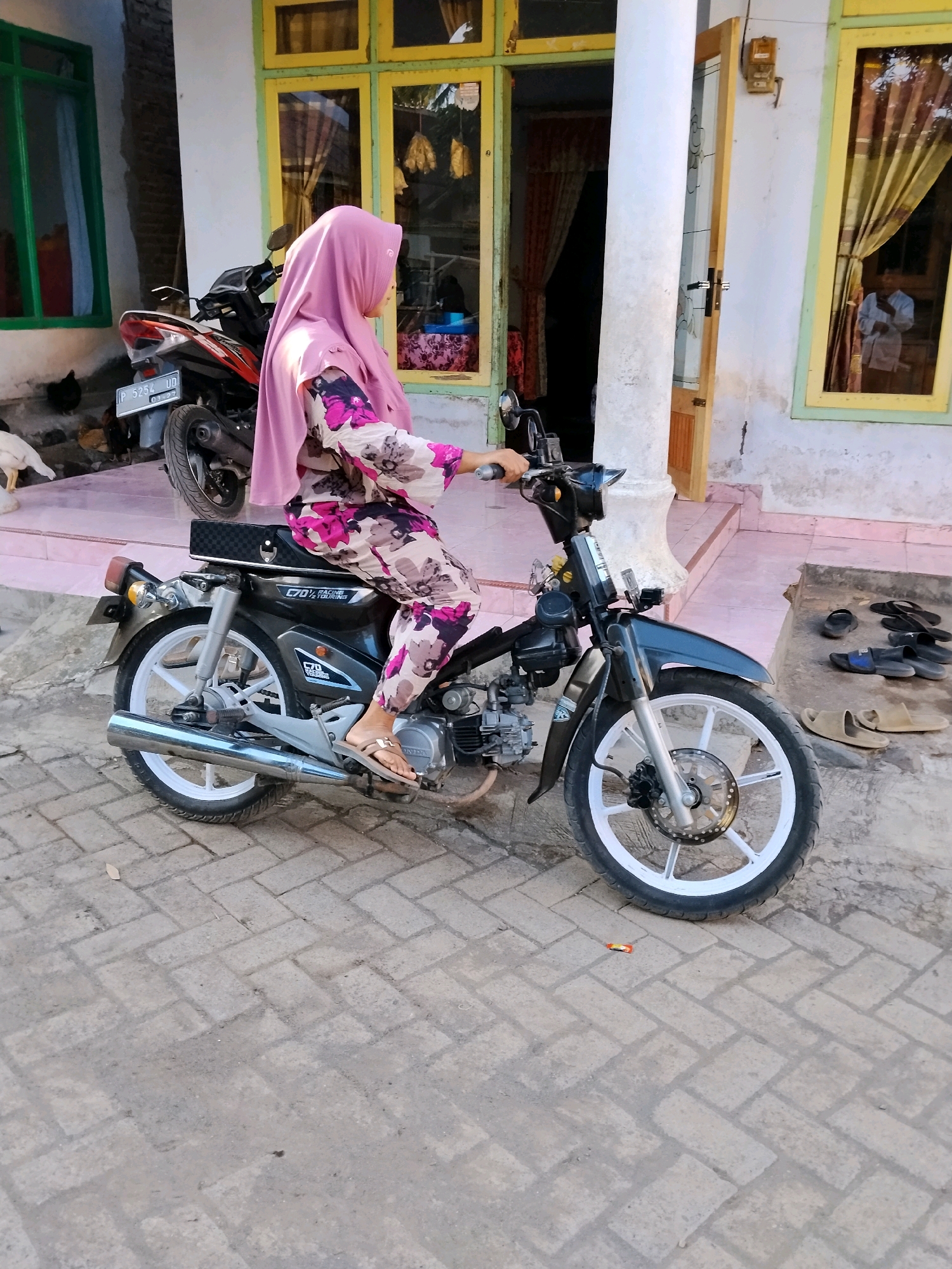 In the city of Panggungan, Indonesia, a young woman wearing a pink hijab and a floral-patterned outfit is seated on a black motorcycle. Her left hand is on the handlebar, suggesting she is preparing to ride. The motorcycle is parked on a patterned brick surface in front of a building with yellow-trimmed windows and a white pillar. Behind her, another motorcycle is visible. The environment appears to be a residential area.