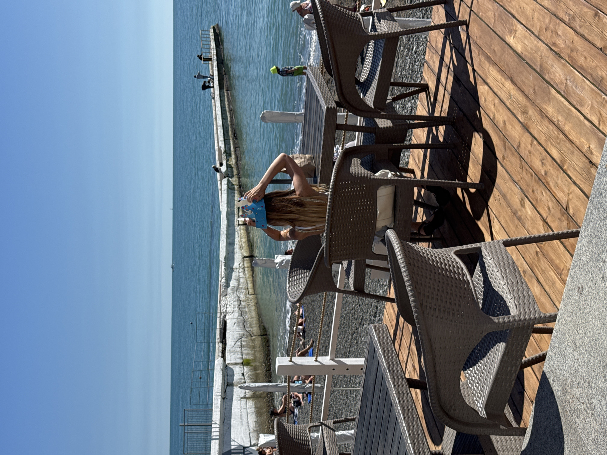 The image captures a vibrant beachside scene, likely from a cafe or restaurant terrace, overlooking the Black Sea in Sochi, Russia. The view is dominated by a clear blue sky meeting a calm, equally blue sea on the horizon.
In the foreground, a wooden deck with dark brown wicker-style chairs and tables suggests an outdoor dining or lounge area. Strong, defined shadows cast by the chairs and tables indicate bright mid-day or early afternoon sunlight. A person, identified as a woman with long blonde hair, is seated at one of these tables, her back to the camera. She appears to be looking down at a light blue object, possibly a phone or tablet with a decorative cover, held in her hands. Her arm is raised as she interacts with the device.
Beyond the wooden deck and a pebbled beach, a concrete pier or breakwater extends into the sea. Several individuals are scattered along this structure. Closer to the foreground, a person in bright green attire stands near the water's edge by the pier. Further along the pier, other people are visible, some standing, some possibly fishing or observing the water. In the far distance, a small boat can be faintly seen on the horizon. The overall impression is one of leisure and relaxation on a sunny day by the sea. No specific text is clearly visible in the image.