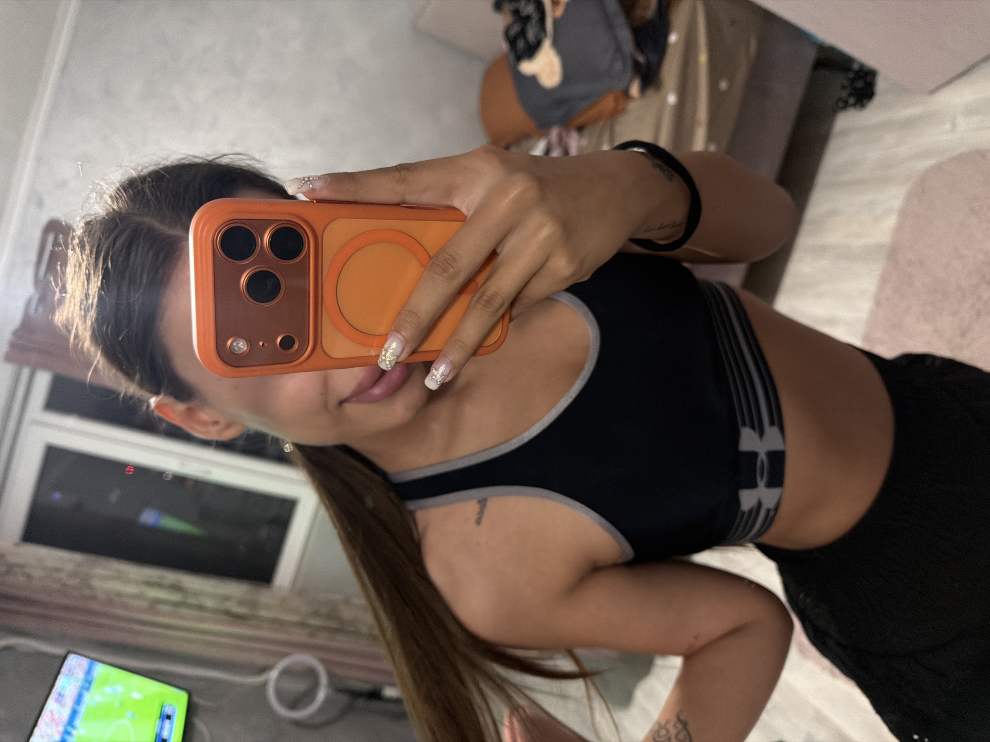 A young woman with long brown hair tied in a ponytail takes a selfie in a room. She is wearing a black sports bra with grey trim and black shorts. Her right hand is holding an orange phone case with a black phone inside, showing the back camera lenses. Her left hand is visible, displaying manicured nails with white glitter and a white floral design. She has a tattoo on her left shoulder and another on her right forearm. In the background, a television screen displays a bright green football field. The room appears to be a bedroom, with bedding visible and a blurry object on a surface to the right. The environment suggests an indoor residential setting. There is no indication of the city of Burgas, Bulgaria.