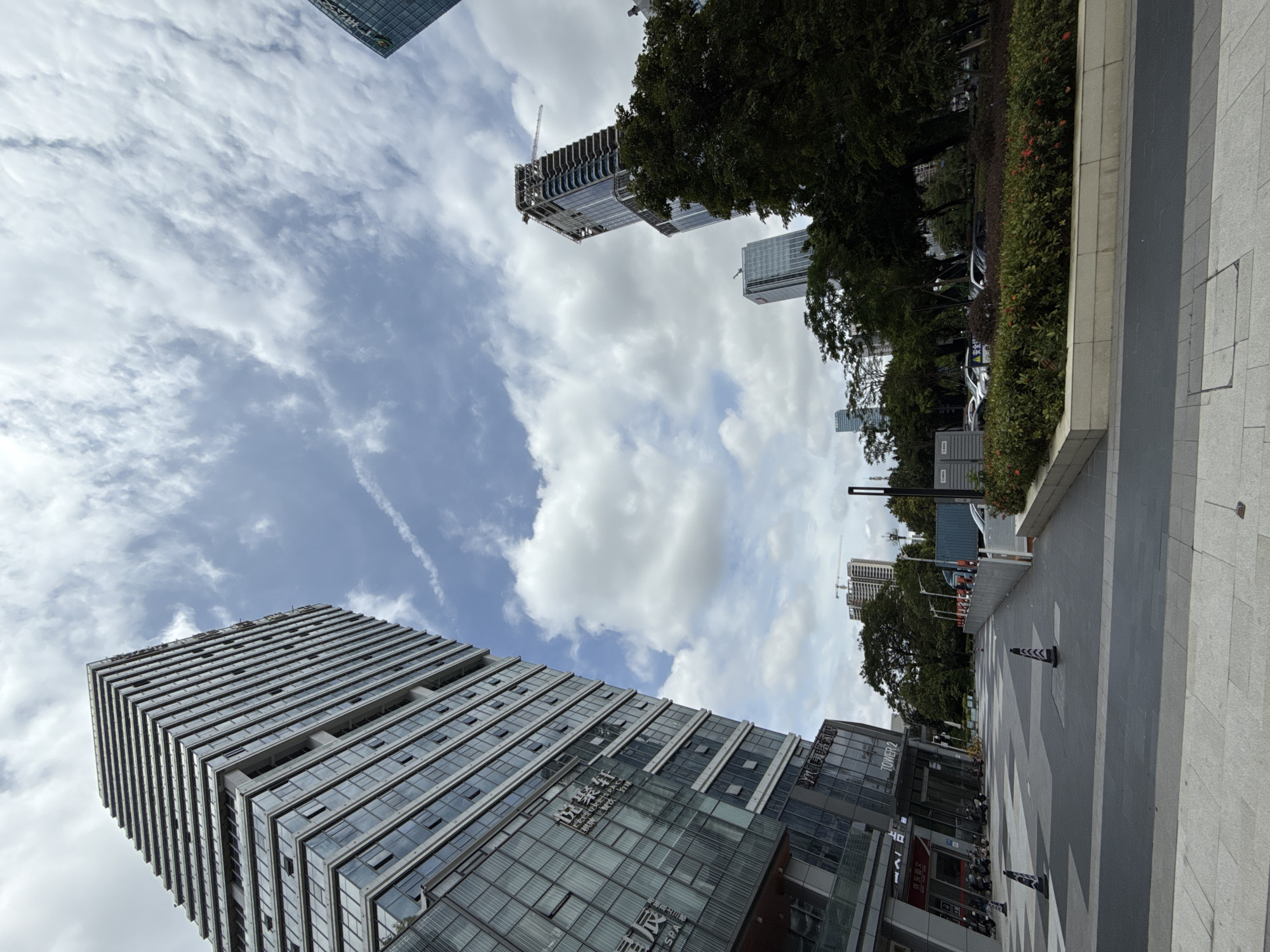 A low-angle, wide shot captures a modern urban scene in Huangpu District, China, dominated by sleek, glass-facaded skyscrapers and a cloudy sky. In the foreground, a paved walkway with a geometric pattern leads towards a row of buildings. To the left, a tall, stepped skyscraper with a facade of glass and grey concrete stands prominently. Its lower levels feature storefronts, one of which displays Chinese characters and the words "TOWER 2" and "SPA". Several parked motorcycles and bicycles are visible near the entrance of this building. Further down the walkway, traffic cones with reflective stripes mark a section of the path.
Beyond the foreground, a lush green park area with trees and red flowering bushes separates the walkway from a street where several cars are parked. In the middle ground, other modern buildings, some appearing to be under construction with cranes visible, rise against the sky. The sky itself is a dynamic mix of bright blue and scattered white cumulus clouds, with a faint contrail etched across it. The overall impression is one of contemporary urban development with pockets of greenery.