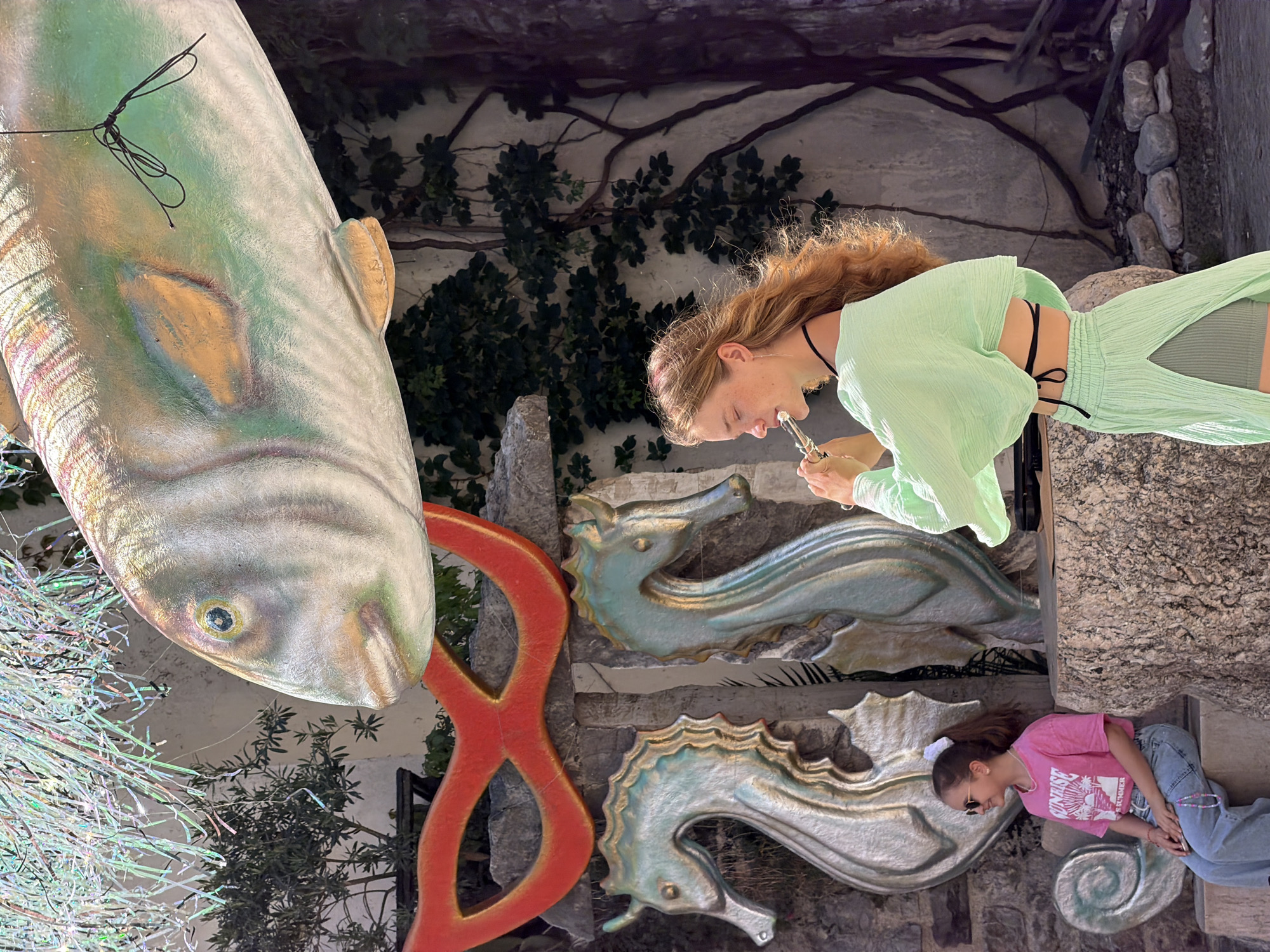 The image shows an outdoor scene, likely in Budva, Montenegro, as evidenced by the distinct architectural style. A woman with red hair, dressed in a light green top and pants set with cutouts, is holding a small silver instrument to her lips. Below her, another woman in a pink t-shirt and jeans is lying on a stone structure, wearing sunglasses.
The background features a textured stone wall with ivy and branches. In the foreground, a large artificial fish hangs, alongside a red figure-eight sculpture. Relief sculptures of sea creatures are affixed to the stone wall. The image has been rotated counter-clockwise.