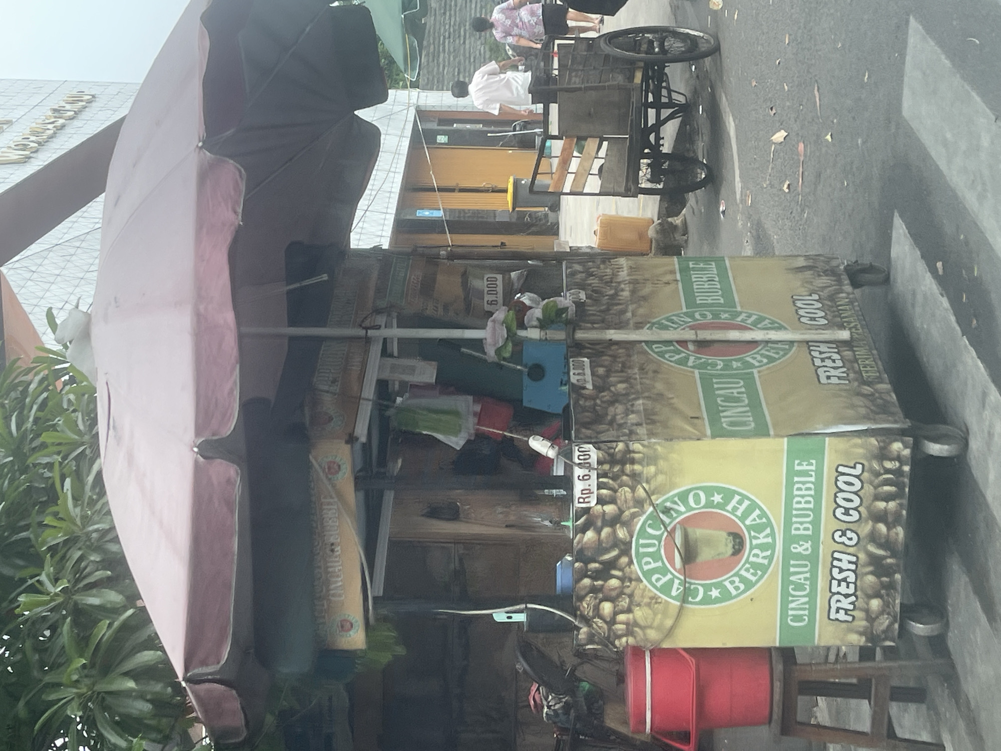 The image depicts a street scene in West Jakarta, Indonesia, focusing on a mobile food cart selling "Cincau & Bubble." The cart is adorned with graphics of coffee beans and a drink, with prices like "Rp. 6.000" displayed. To the left of the cart, a red bucket sits on a wooden stool. In the background, two individuals, a man in a white shirt and a woman in a floral shirt, walk away from the camera. A horse-drawn cart with wooden planks is visible behind them, and a yellow trash can is nearby. The environment appears to be an urban street with buildings and foliage contributing to the city backdrop.