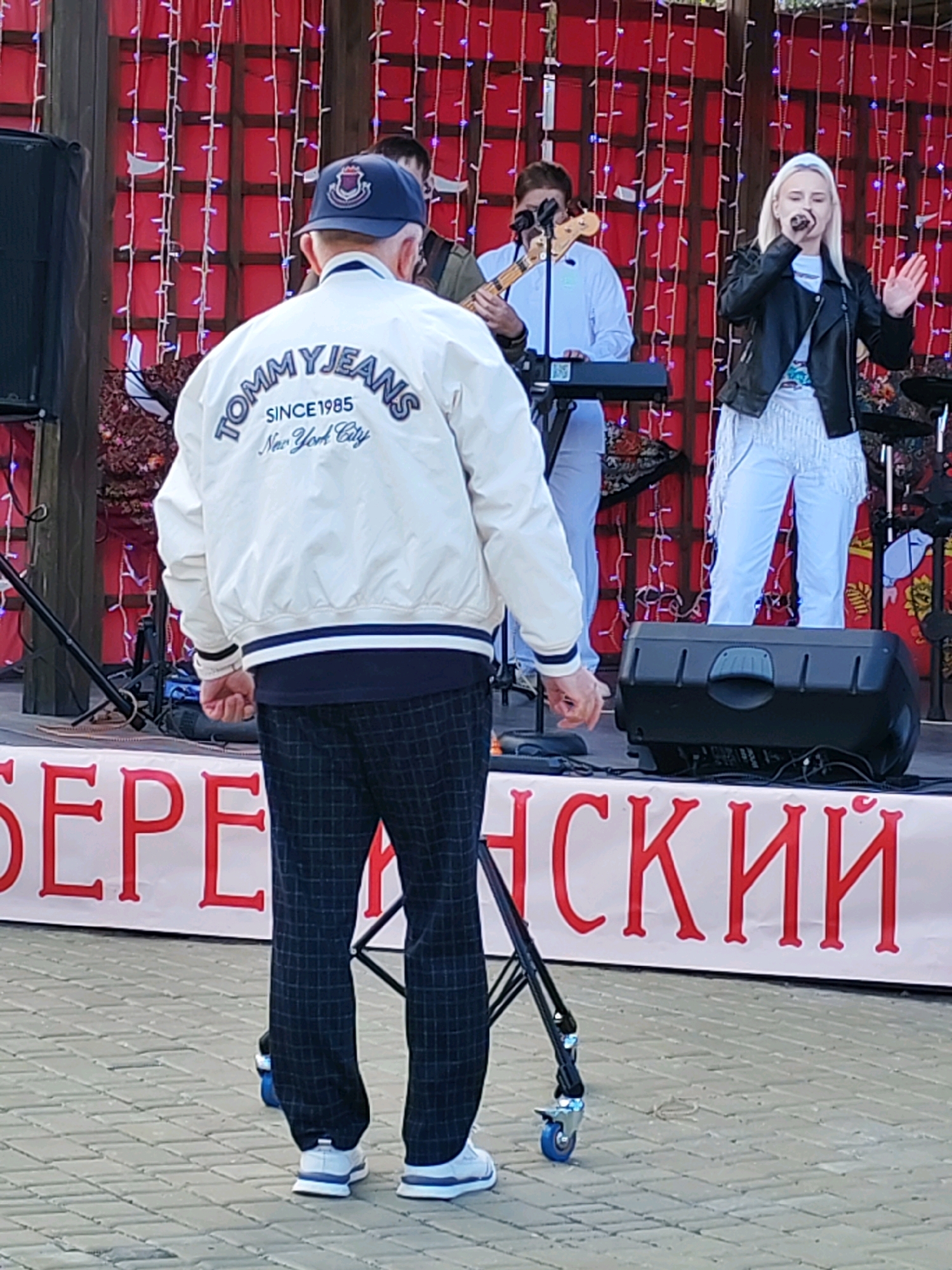 A man stands in the foreground, facing away from the camera, watching a band perform on a stage. He is wearing a white jacket with "TOMMY JEANS SINCE 1985 New York City" printed on the back, a blue baseball cap, and plaid trousers. The band consists of a woman singing and a man playing a bass guitar. The stage is decorated with red fabric and strings of lights. A large black speaker is placed on the stage in front of the band. A banner with red Cyrillic lettering is visible at the bottom of the image. The ground is paved with interlocking grey stones.