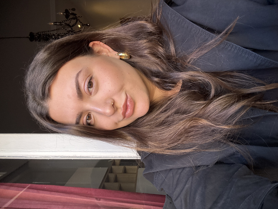 A female subject with long, dark brown wavy hair, brown eyes, a small nose ring on her left nostril, and multiple earrings including a prominent gold hoop in her right ear, is positioned in a head-and-shoulders shot, looking directly at the camera with a slight smile. She is wearing a dark, possibly black, long-sleeved top or jacket. The scene is set indoors in a building located in Madrid, Spain. To the subject's left, a white vertical architectural element, likely a window frame, is visible. Beyond it, a reddish-brown curtain partially obscures a brightly lit background featuring a grid-like shelving unit. To the subject's right, a darker interior space reveals a prominent black, ornate chandelier hanging from the ceiling. Natural light appears to originate from the left side of the frame, illuminating the subject's face and creating some shadow on her right side.