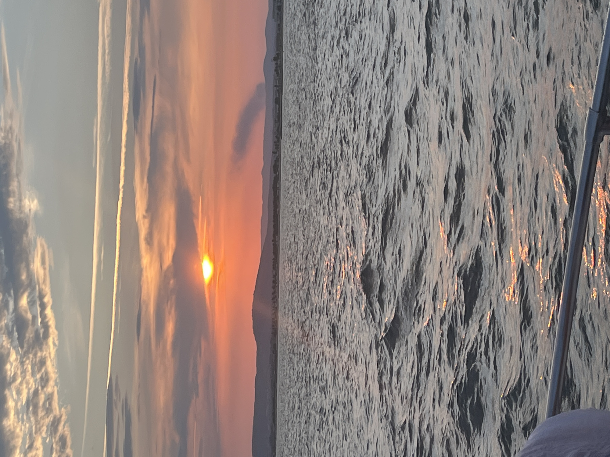 This is a photograph of a sunset over the Mediterranean Sea, taken from a boat in Palavas-les-Flots, France. The sun, a bright orange orb, is low on the horizon, partially obscured by pink and orange clouds. The sea is choppy, with whitecaps reflecting the sunset's light. In the distance, a hazy silhouette of hills and coastline can be seen. A plume of dark smoke rises from the land in the middle distance. The foreground shows a metal railing and a person's shoulder, indicating the vantage point is from a boat.