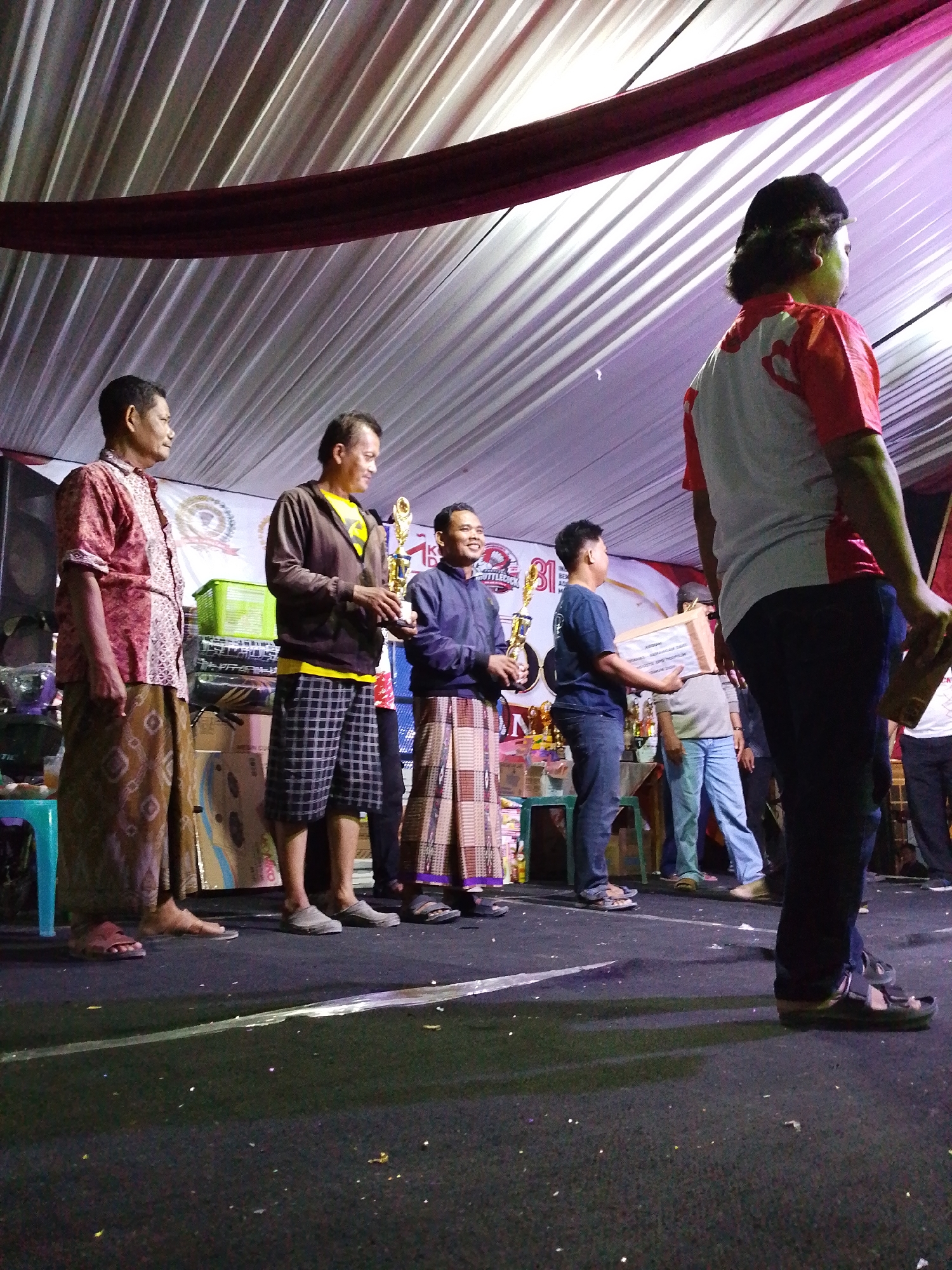The image shows a group of men standing on a stage, possibly at an awards ceremony or event, under a white tent with purple accents. The men are dressed in traditional attire, including sarongs. One man in the center is holding a trophy. Another man to his right is holding a framed certificate or award. In the background, there are other people, trophies, and a banner. The stage is covered in black fabric, with some confetti scattered on it. The lighting suggests an indoor or evening event. The event appears to be taking place in Lawatan, Indonesia, as indicated by the banner in the background.
