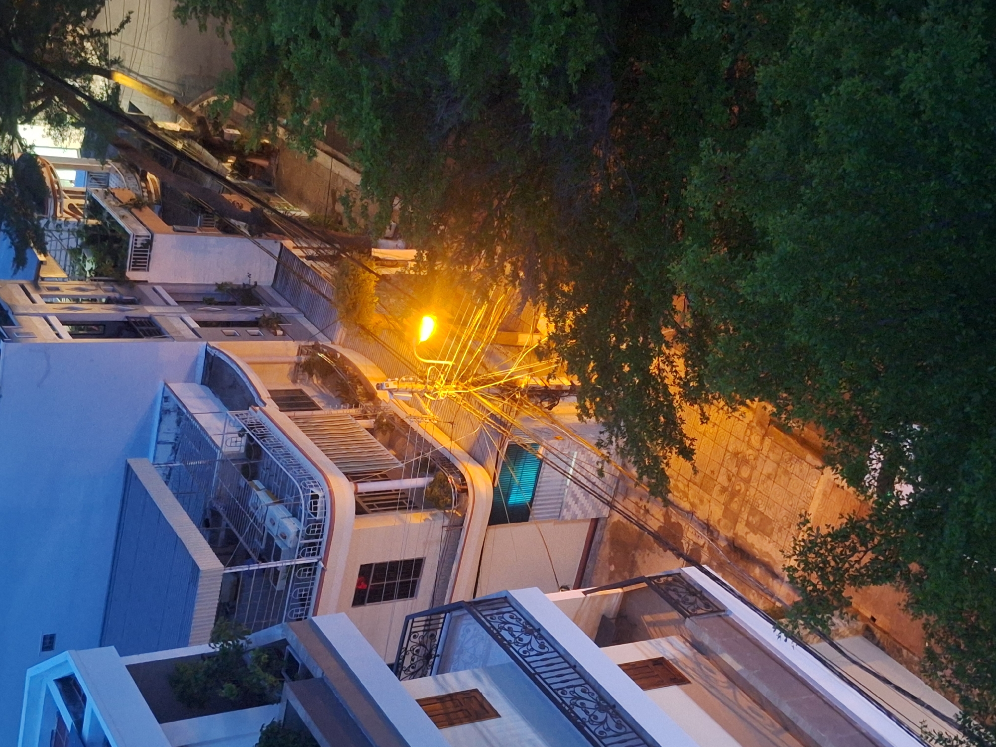 This aerial shot captures a street scene in Nha Trang, Vietnam, during dusk or early night. A bright street lamp illuminates a tangle of electrical wires overhead, casting a warm glow on the textured pavement below. Flanking the street are multi-story buildings with balconies, windows, and outdoor areas, some featuring architectural details like railings and awnings. Lush green trees are interspersed throughout the scene, their foliage obscuring parts of the buildings and the sky. The overall impression is one of a residential or commercial street in a Southeast Asian city transitioning from day to night, with the man-made structures contrasting with the natural elements.