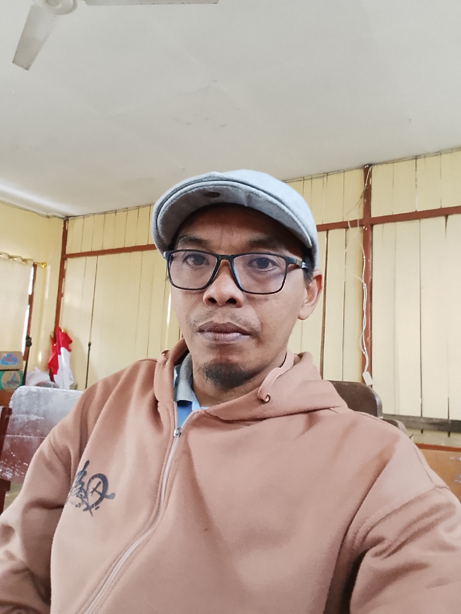 A medium shot shows a man in a light brown hooded sweatshirt, a gray flat cap, and black-rimmed glasses. The man is looking directly at the camera. The background appears to be an indoor room with yellow walls and a white ceiling with a fan. There are blinds on the wall behind him, and a red and white flag is visible to the left of the frame. The man is wearing a blue collared shirt under his sweatshirt.