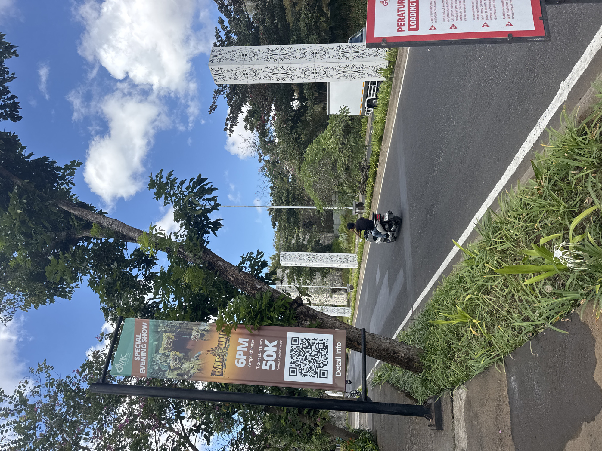 In Kuta Selatan, Indonesia, a street scene unfolds under a partly cloudy sky. A tall, dark lamppost stands on the left, supporting a banner advertising a "SPECIAL EVENING SHOW" featuring "BARONG." The banner, in shades of green and brown, displays graphics of traditional masks and text indicating show times, ticket prices, and a QR code for more information. A mature tree with green foliage grows near the lamppost.
The road in front of the banner is asphalt, marked with a white line. A person rides a white scooter away from the viewer on the right side of the road. On the right side of the road, a white truck is parked, partially visible. Also on the right, two tall, decorative white pillars with intricate patterns stand along the roadside. Adjacent to the road, lush green plants and a white flower with thin petals are visible. A red and white sign is partially visible on the far right. The environment appears to be an outdoor public space, likely a tourist area or entertainment venue.