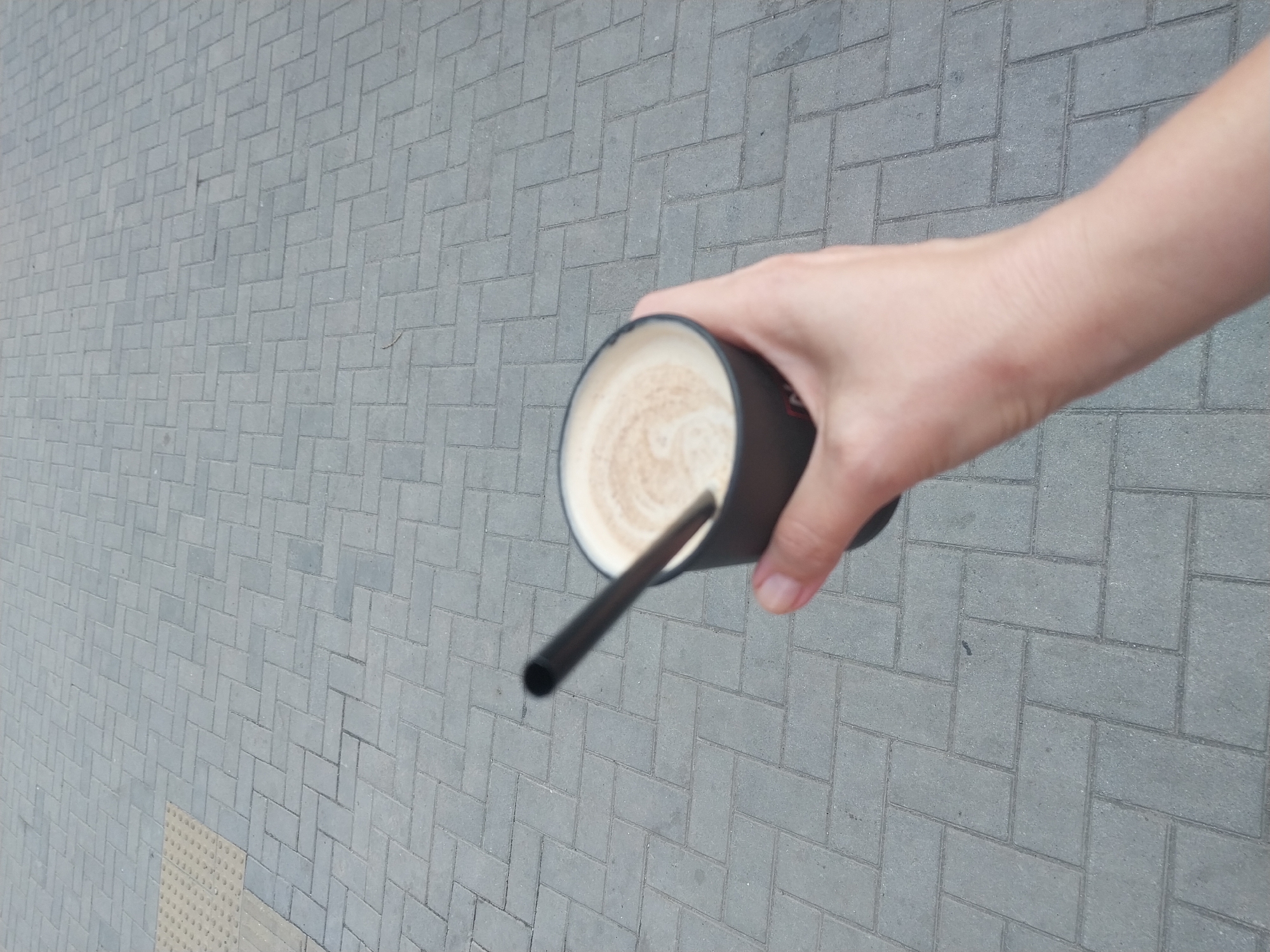 In Dnipro, Ukraine, a person's hand is holding a black paper cup filled with a frothy beverage, possibly coffee with milk, with a dark straw inserted. The cup is black and has some white lettering on the side. The hand, with visible skin texture, is positioned in the lower right quadrant of the image. The background consists of gray interlocking paving stones, characteristic of city sidewalks. A raised tactile paving surface, typically for visually impaired individuals, is visible in the upper left corner, suggesting an urban pedestrian area. The lighting indicates it is daytime.