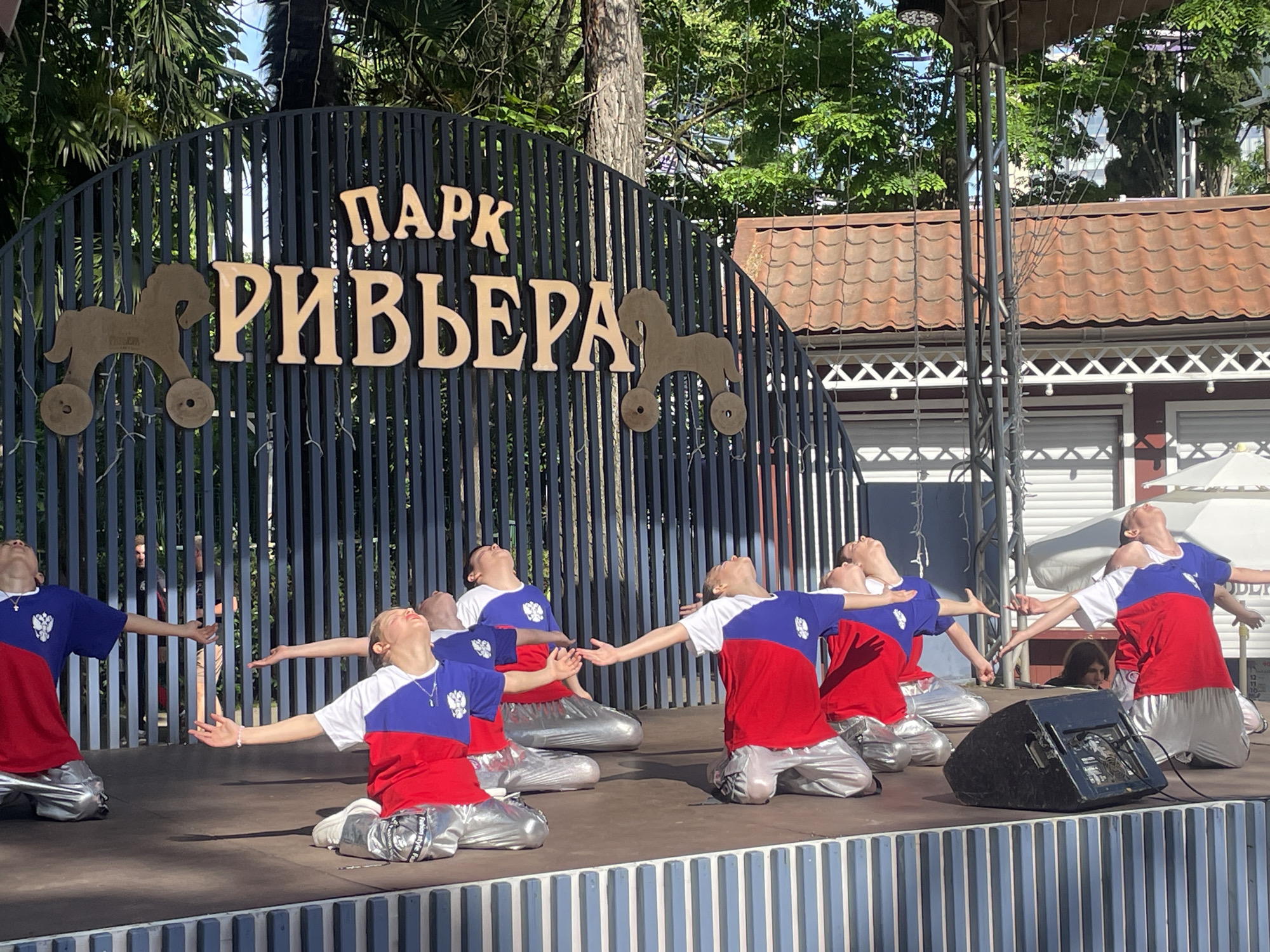 In this medium shot photograph taken in Sochi, Russia, a group of children in matching outfits perform a synchronized dance on a wooden stage. The stage is set in front of a dark metal fence adorned with the words "ПАРК РИВЬЕРА" (Riviera Park) in large, light-colored letters. Wooden horse figures are affixed to the fence on either side of the park's name. The children are all wearing bright red, white, and blue shirts with a crest on them, and shiny silver pants. They are kneeling on the stage, looking upwards with their arms extended. The background reveals lush green trees, a tiled roof structure, and some fencing, suggesting an outdoor park setting. The lighting indicates it is daytime.