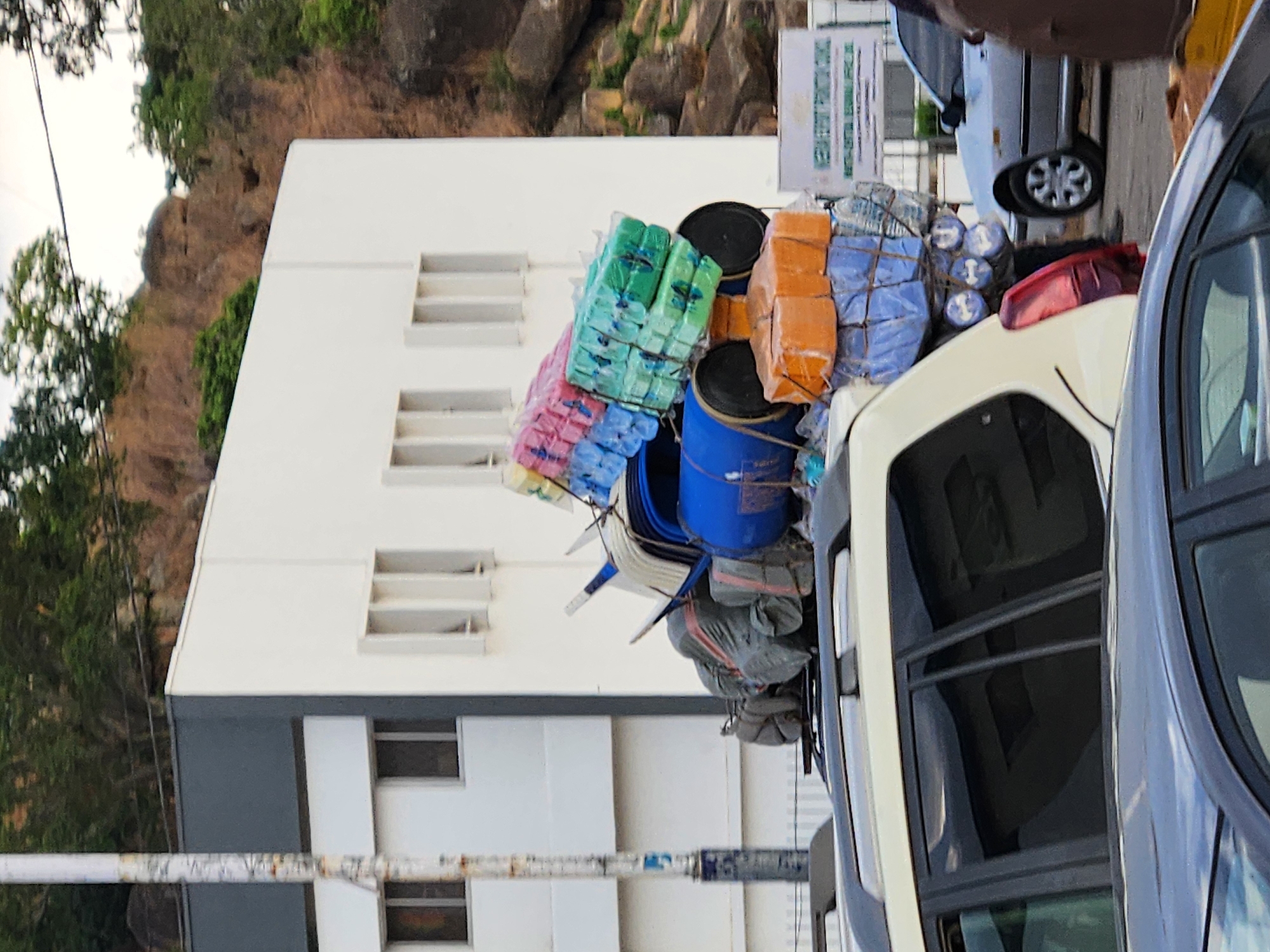 The image shows a vehicle heavily loaded with various items, including packaged goods, barrels, bags, and chairs. These items are secured on the roof of the vehicle with rope. The background consists of a white and grey building with several windows. In the distance, there is a hill with rocks and trees. The scene is captured in the city of Unknown, Unknown. Another car is partly visible in the foreground, suggesting the presence of traffic or a parked vehicle. The overall impression is one of a congested urban environment with possibly overloaded transportation.
