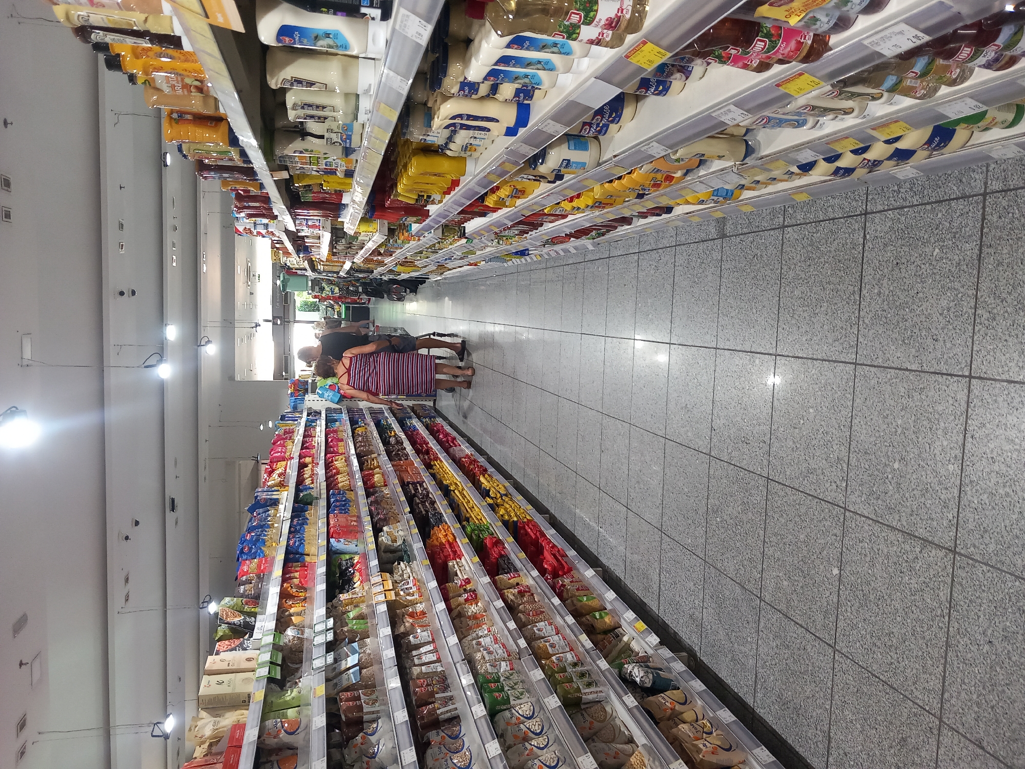 The image shows an aisle in a supermarket in Kos, Greece, filled with shelves of food products. The shelves are stocked with various packaged goods, including bags of pasta, rice, snacks, and bottles of sauces and condiments. Two people, a man and a woman, are walking down the aisle away from the camera, facing the shelves. The woman is wearing a striped dress, and the man is wearing a dark t-shirt and shorts. The supermarket has tiled flooring and fluorescent lighting overhead. The overall scene depicts a typical grocery shopping environment.