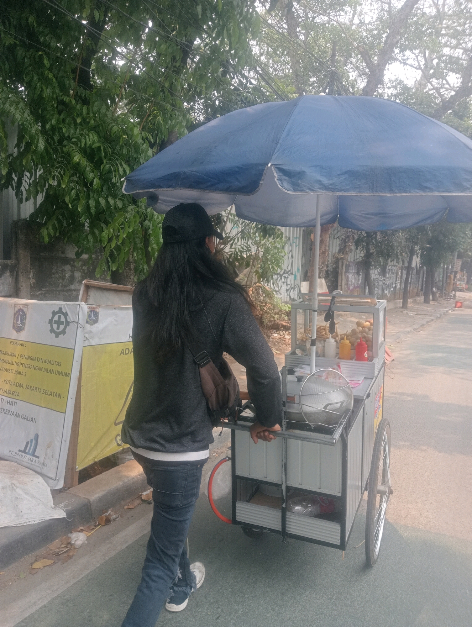 An individual wearing a black cap and dark-colored hoodie pushes a food cart with a blue umbrella on a street in South Jakarta, Indonesia. The food cart is equipped with a glass display case containing various food items, bottles of condiments, and a metal sieve. The cart has silver and grey metal siding and two wheels, one of which is visible on the right side of the image. The individual is seen from behind, walking away from the camera. The background consists of trees, a concrete wall with graffiti, and a road. A large banner with text and graphics is positioned to the left of the individual. The scene suggests a street food vendor operating on a sunny day.