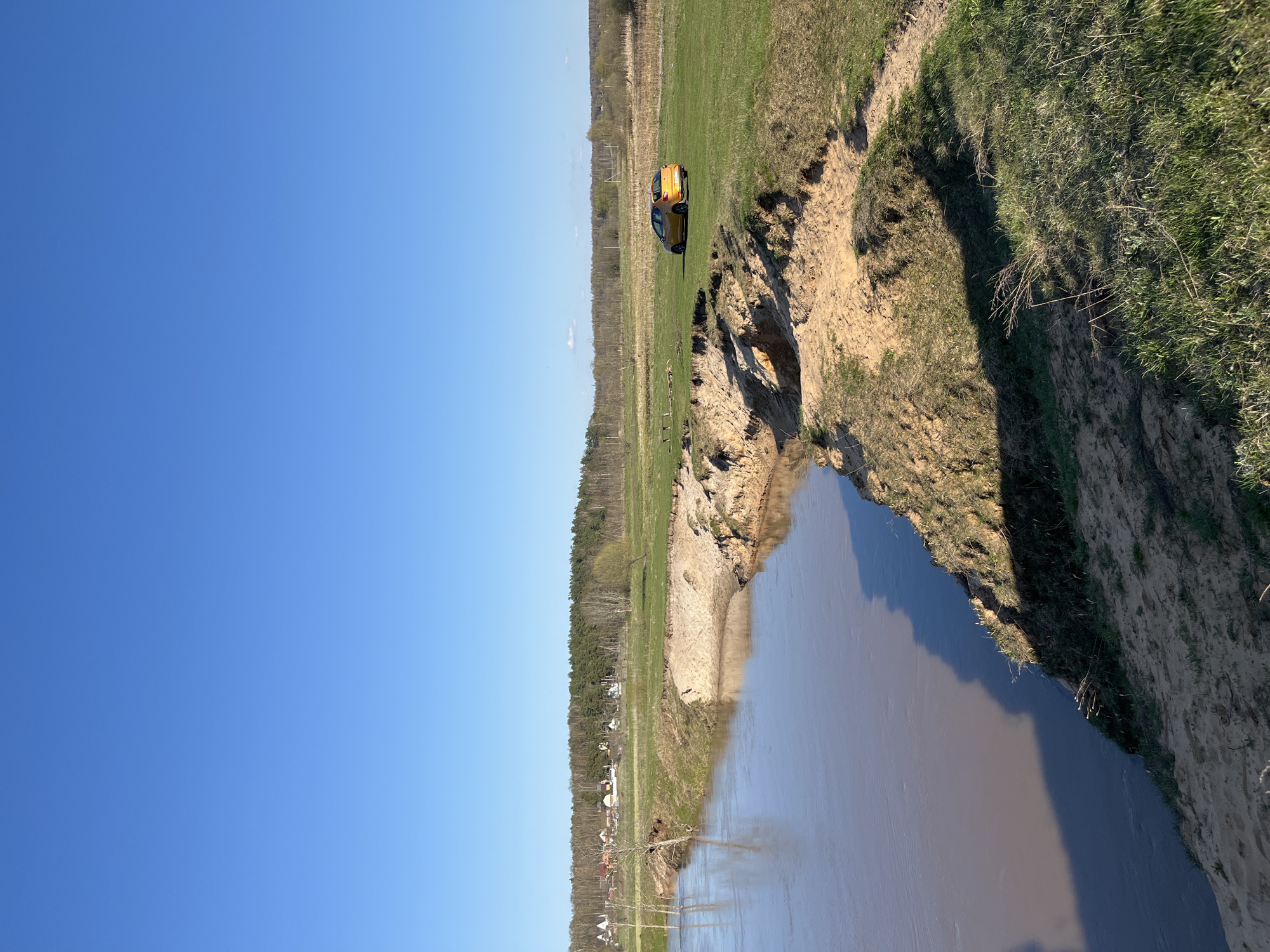 The image shows a body of water with a steep, sandy bank on the right side, and trees and a small village in the distance on the left side. In the center of the frame, an orange car is parked on a grassy area near the edge of the bank. The sky is clear and blue. The scene is located in the city of Voronino, Russia.
