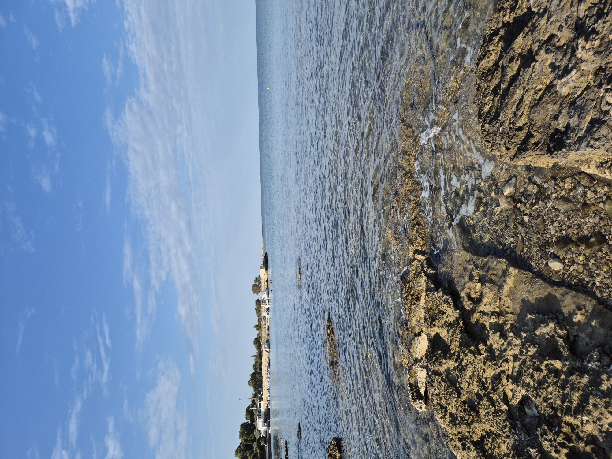 The image depicts a tranquil coastal scene in Zambratija, Croatia. The foreground is dominated by large, weathered rocks and pebbles, with the clear, shallow water gently lapping against them. Small waves create foamy white lines as they recede from the shore.
In the middle ground, the calm sea extends towards a harbor visible in the distance. Several small boats are docked, and a few buildings line the shore, backed by lush green trees. The sky above is a brilliant blue, adorned with scattered white clouds, suggesting fair weather. The horizon is sharp, with the sea meeting the sky in a clean line. A small yellow buoy is visible in the far distance on the water. There are at least two people visible in the water near the harbor.