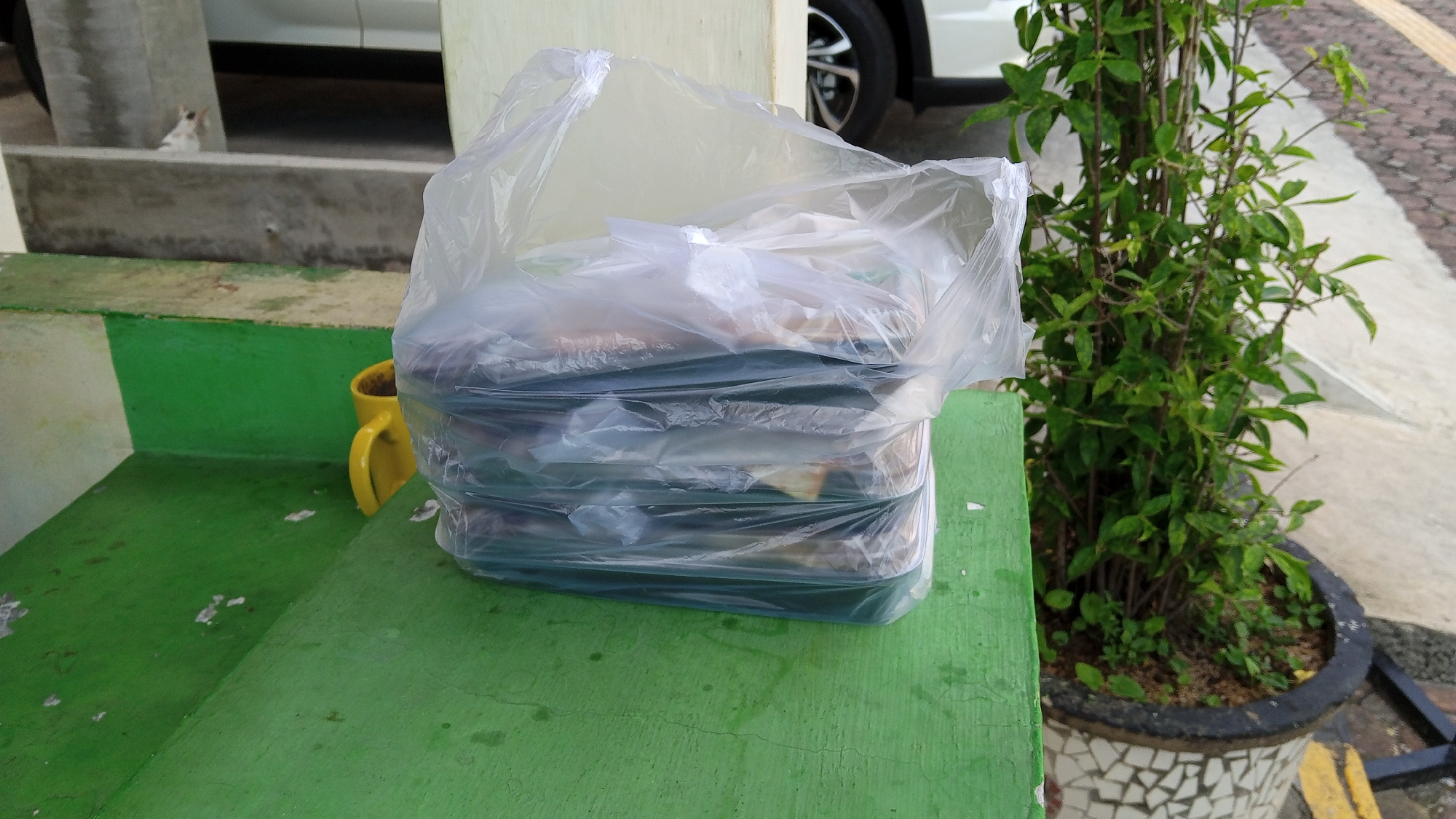 A stack of food containers, wrapped in a clear plastic bag, sits on a green-painted surface. To the left of the containers, a yellow mug is visible. In the background, a white car and a potted plant with green leaves are present. The scene appears to be outdoors, possibly on a public step or ledge. The surrounding environment is a city in Bireuen, Indonesia, with hints of pavement and concrete structures.