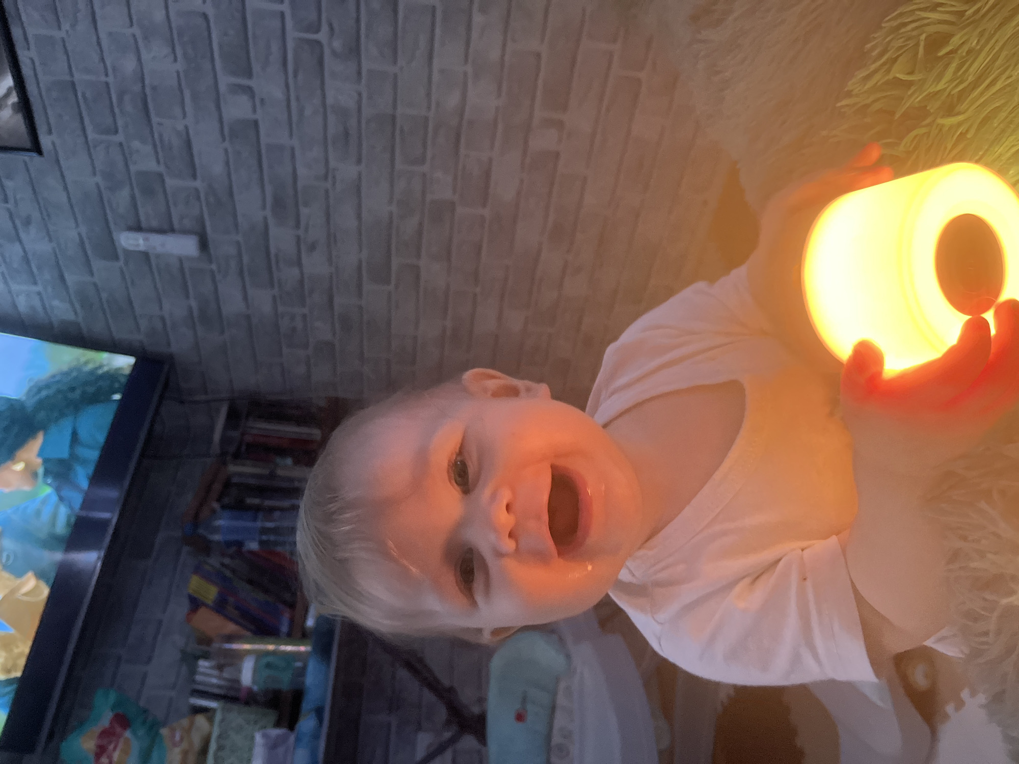 A close-up, slightly rotated portrait shot shows a fair-skinned baby in a white shirt holding a yellow light in their hands. The baby is smiling with their mouth open, revealing their tongue. The background includes a brick wall, a shelf filled with books and objects, and a television displaying an animated image. The baby's hands glow red from the light they are holding. There is no information to suggest that this photo was taken in Vesyoloye, Russia.