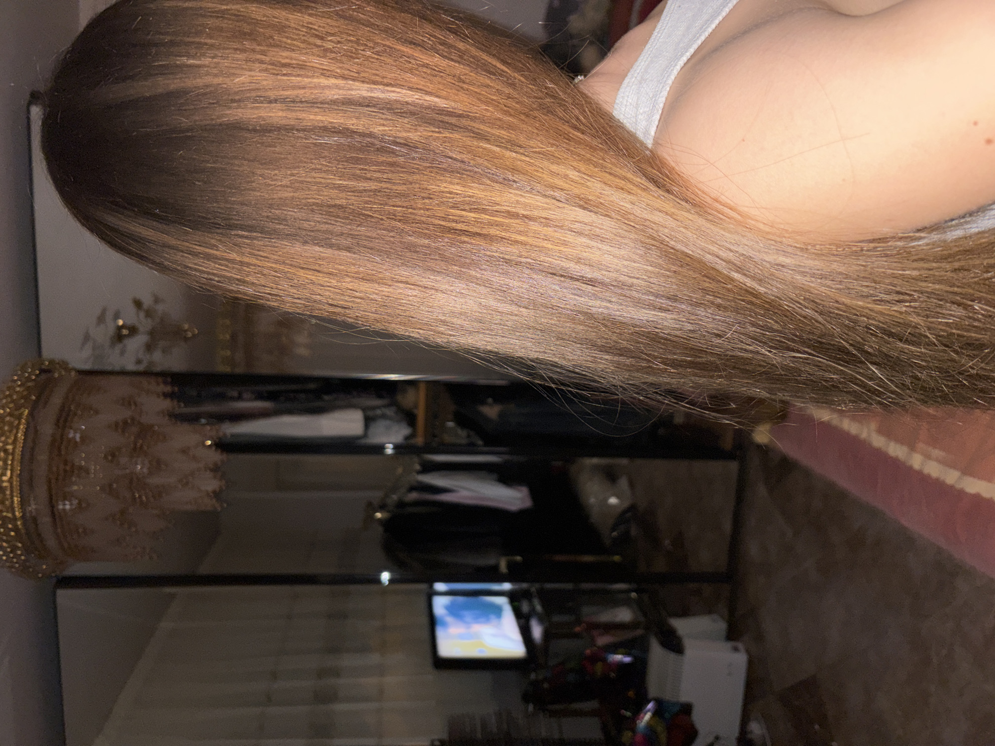 The image shows a person with long, light brown hair. The person is wearing a white tank top. Behind the person is a closet with shelves. A TV is visible in the closet, displaying a bright blue screen. There is also a decorative gold crown on top of the closet. The location is most likely indoors in a private residence. There is no evidence to suggest this image was taken in Sofia, Bulgaria.