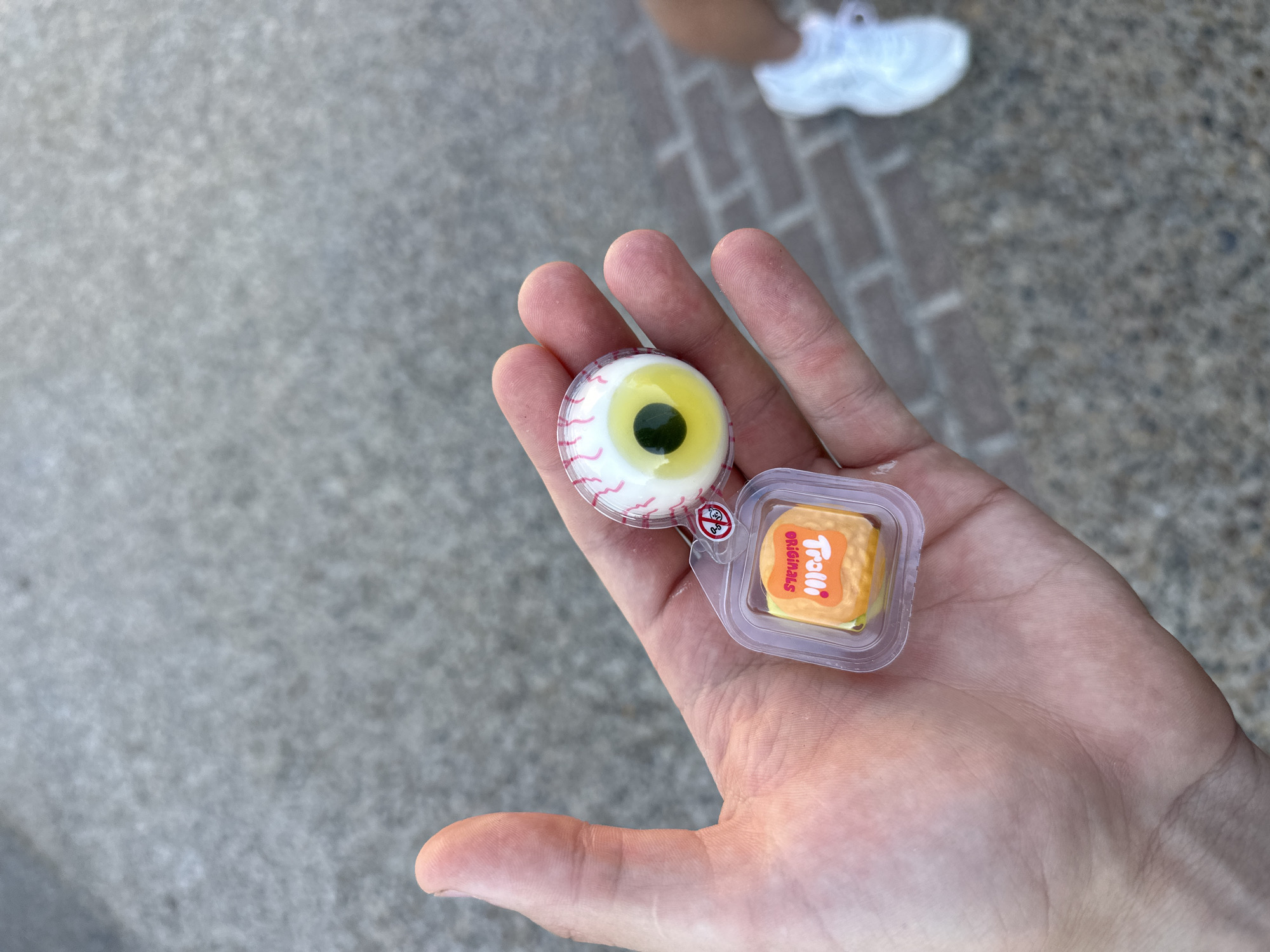 In this close-up shot, a person's hand is held up, displaying two small candies. The hand is positioned in the foreground, with the palm facing upwards, and fingers slightly curled. The skin tone of the hand suggests it belongs to a fair-skinned individual.
The first candy, on the left, is designed to look like a human eyeball. It has a white outer layer with red veins, a yellow iris, and a dark green pupil. It appears to be a gummy candy.
To the right of the eyeball candy is a small, clear plastic container holding a single, square-shaped candy. This candy is orange and yellow and has the "Trolli Originals" logo printed on it in red lettering. The packaging is typical for individual candy servings.
The background is a blurred view of a paved surface, likely a sidewalk or pathway, composed of greyish concrete and brick-like patterns. In the upper right portion of the image, a blurred white athletic shoe is visible, suggesting the person is standing or walking outdoors.
The lighting indicates it is daytime, with the sun casting some light on the hand and candies. There are no strong shadows, suggesting overcast conditions or indirect sunlight. The overall scene appears casual and unhurried, possibly during a walk or outing. The focus is entirely on the candies and the hand holding them.