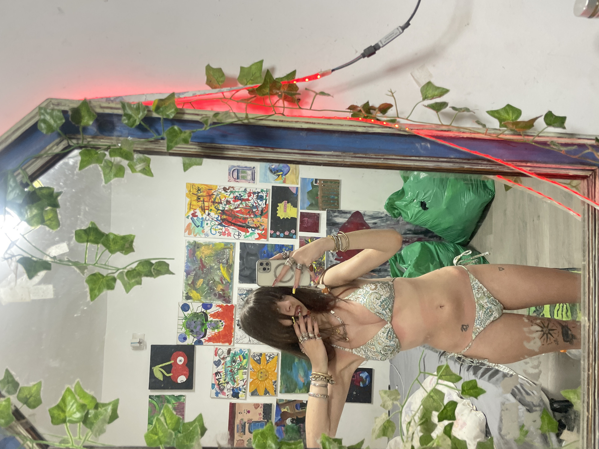 The image shows a woman reflected in a mirror. She is posing for a selfie in a bikini with a paisley print. The background features a wall adorned with colorful artwork. The mirror itself is decorated with artificial ivy and red LED strip lights. A green plastic bag is visible in the background. Location information (Raanana, Israel) cannot be verified from the image content alone.