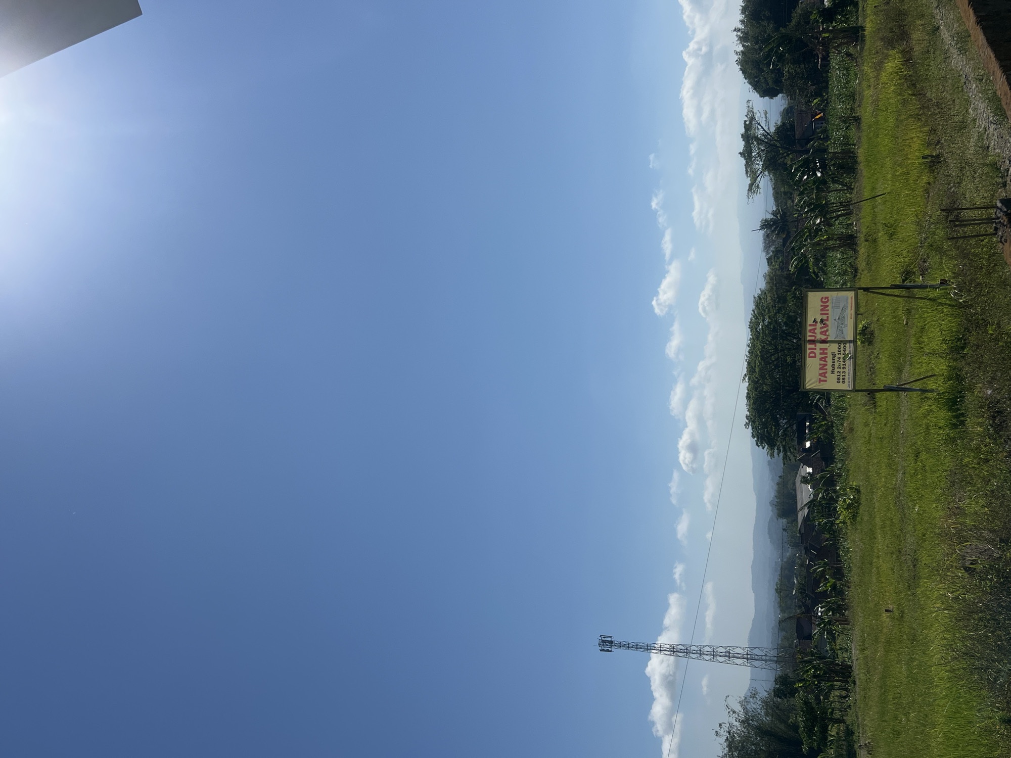 This image shows a bright, sunny day in what appears to be a rural or semi-rural area, likely in Danupayan, Indonesia. The sky is a clear, vibrant blue with a few scattered white clouds. In the foreground, there is a lush green grassy field.

To the left of the image, a tall, slender telecommunications tower stands prominently against the sky. In the mid-ground, there are several houses with various roof types, surrounded by trees and vegetation, including what appear to be banana trees. A yellow sign is visible in the center of the frame, with red text that reads "DIJUAL TANAH KAVLING," which translates to "LOTS FOR SALE." The sign also includes contact information. The background features rolling hills or mountains, adding depth to the landscape. The overall impression is one of a developing area with natural beauty.