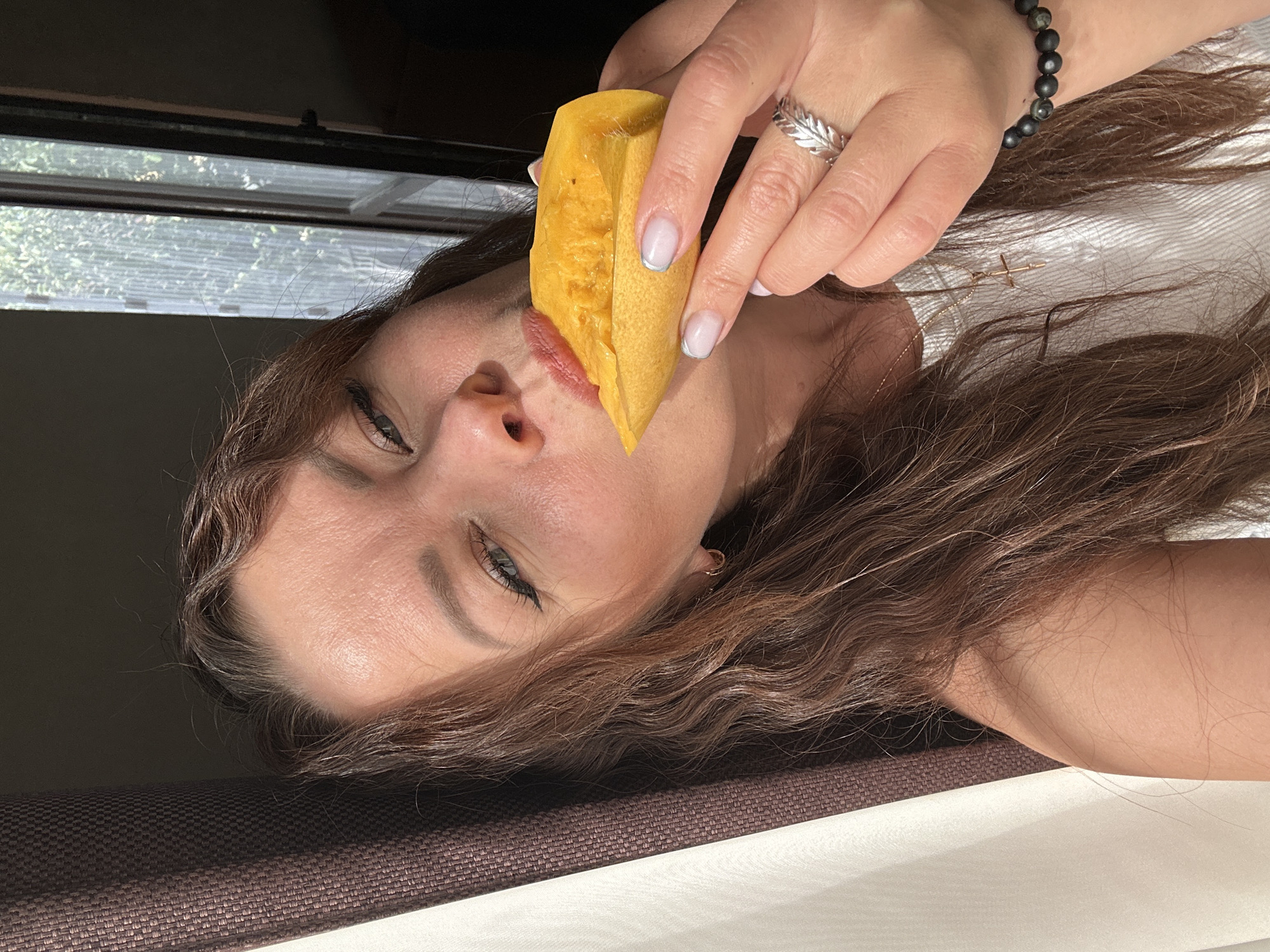 A woman with brown wavy hair is holding a yellow fruit or vegetable slice in front of her mouth. She has fair skin, blue eyes, and is wearing a silver ring and a beaded bracelet. The background includes a window with an outdoor view and a partially visible dark-colored object. The location is unknown, and there is no information to suggest that the image was taken in Chaikovsky, Russia.