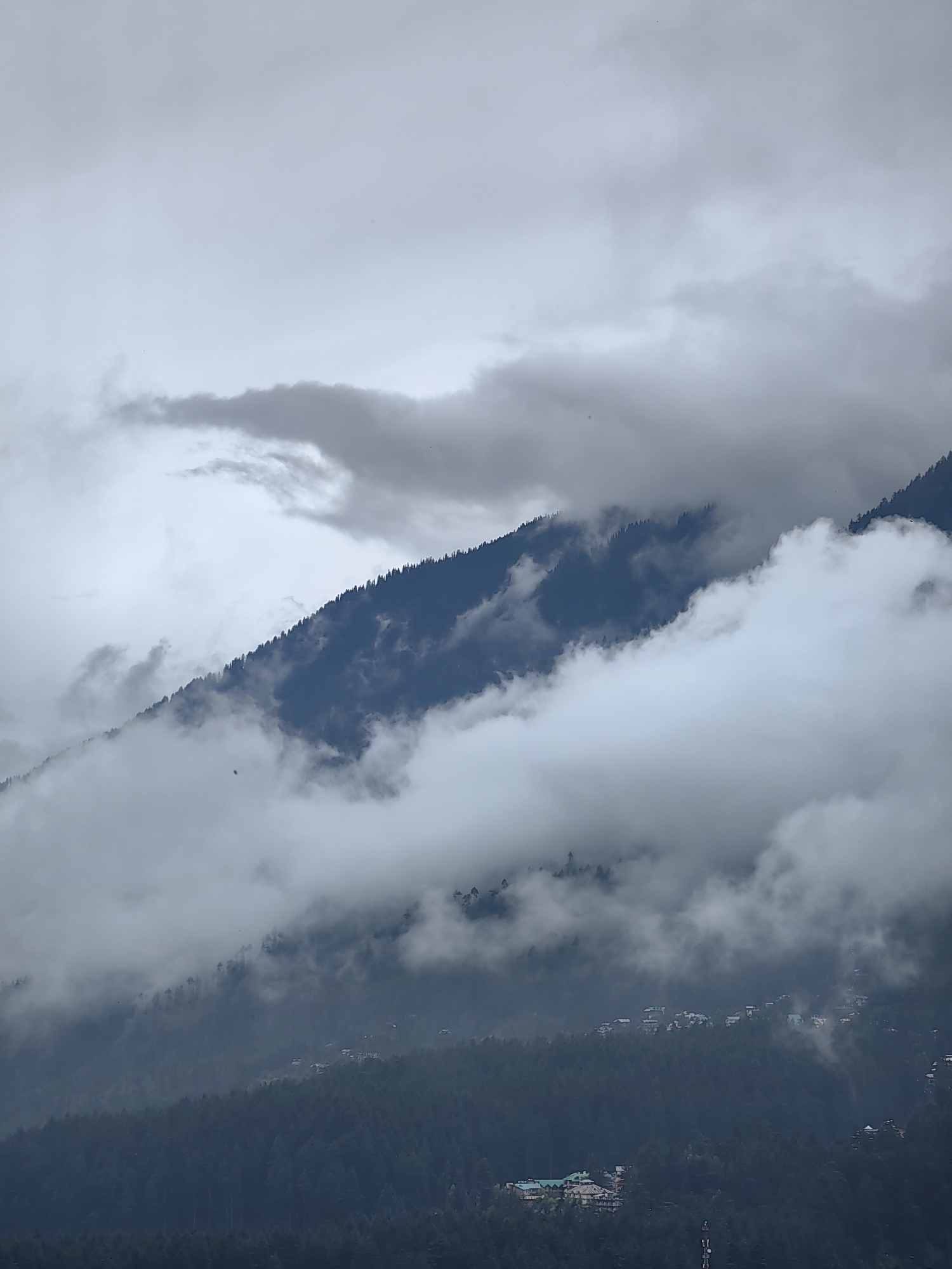 The image shows a mountainous landscape with a forest of pine trees on the slopes. The sky is overcast with thick clouds, some of which are obscuring the mountain peaks and slopes, creating a misty and atmospheric effect. Several buildings, appearing to be houses and a larger structure with a green roof, are scattered across the lower slopes of the mountains, suggesting a village or town. The overall scene depicts a serene, natural environment in a mountainous region.