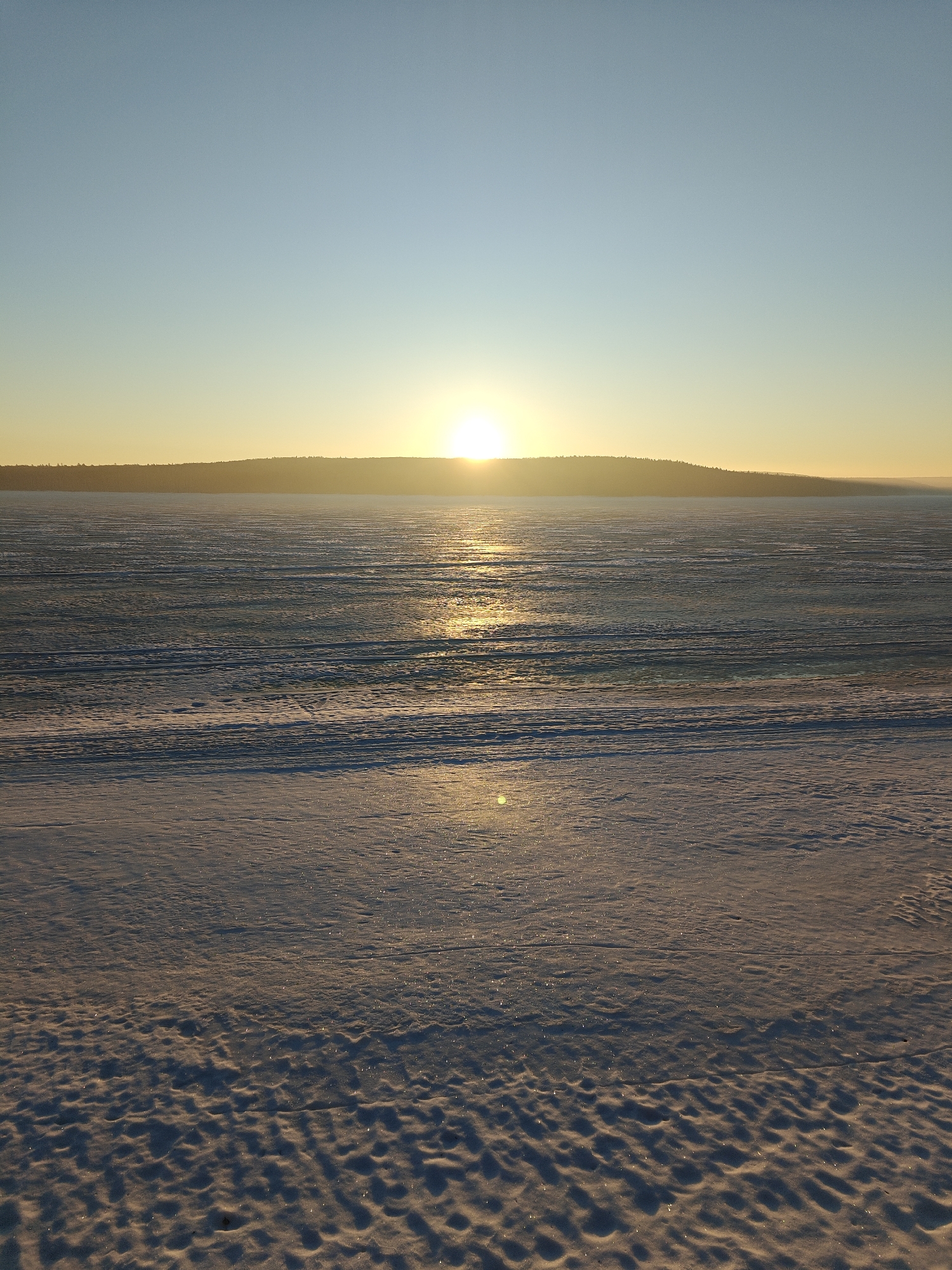 The image depicts a winter landscape near the city of Chaikovsky, Russia, at sunrise or sunset. The sky is a gradient of blue to yellow, indicating the time of day. A distant landmass is visible on the horizon, with the sun positioned slightly above it. The foreground is covered in snow, with textured patterns created by wind or other natural processes. A frozen body of water lies between the landmass and the foreground snow. The ice has visible cracks and ridges, suggesting its frozen state. The sun's reflection is visible on the ice, creating a bright line extending toward the viewer. No people or animals are visible in the scene.