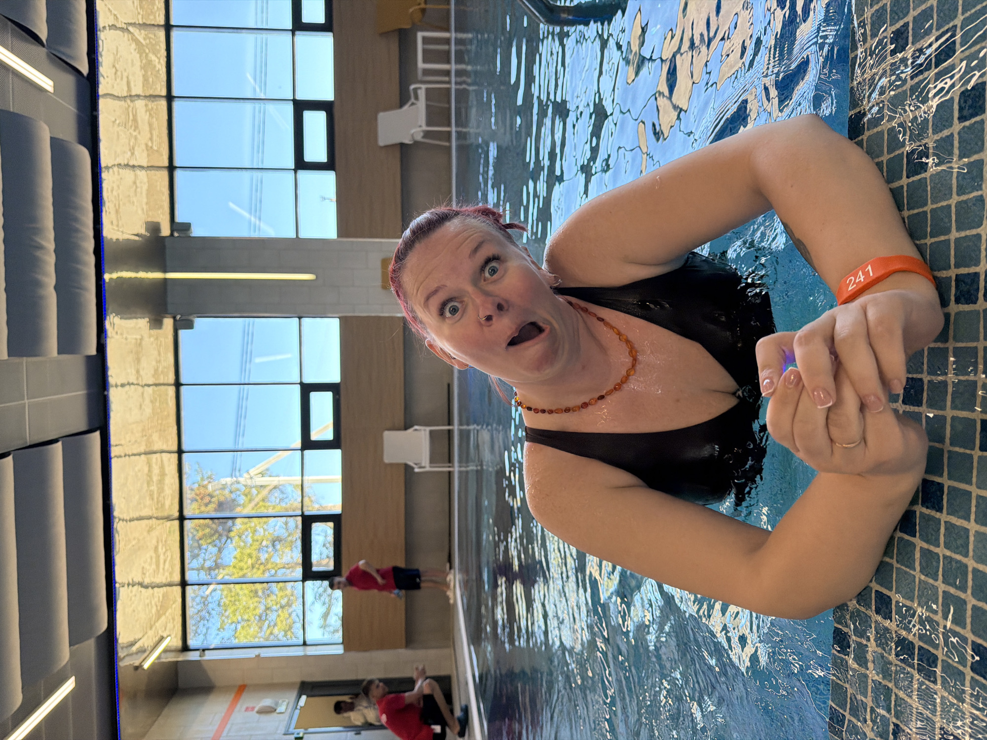 A medium shot depicts a fair-skinned woman in a black swimsuit, perched at the edge of a swimming pool. Her expression is one of wide-eyed surprise, mouth agape. She is wearing a necklace made of amber-colored beads and a wristband with the number 241. The pool's edge is tiled with blue and white squares. In the background, through large windows, are visible a building exterior, trees, and two people in red clothing. Several chairs are visible in the interior background. The location is likely in Votkinsk, Russia, based on additional context.