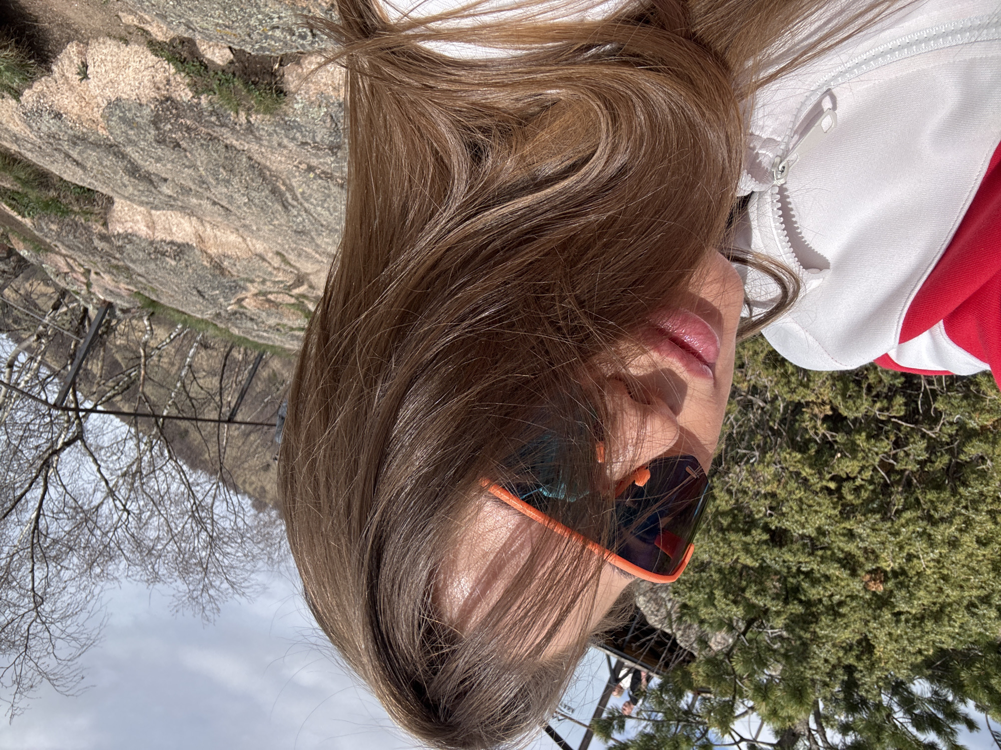 A woman with long brown hair is wearing orange-framed sunglasses and a white and red jacket. She is outdoors, with a rocky cliff and bare trees behind her on the left, and green foliage on the right. The location is Unknown, Russia.