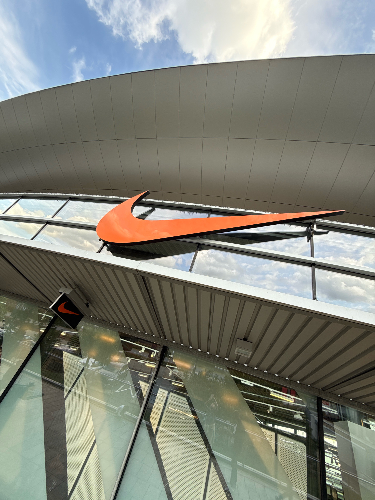 The image shows the exterior of a Nike store in Wolfsburg, Germany. The main subject is a large, orange Nike swoosh logo mounted on the facade of the building. The building features a modern architectural design with a curved, light-colored roof and large glass windows. The windows reflect the sky with scattered clouds. Below the main swoosh, a smaller, illuminated Nike logo is visible on a dark rectangular sign. Inside the store, through the reflective glass, rows of display lighting and shelves are partially visible. The overall impression is a clean, contemporary retail space under a bright sky.