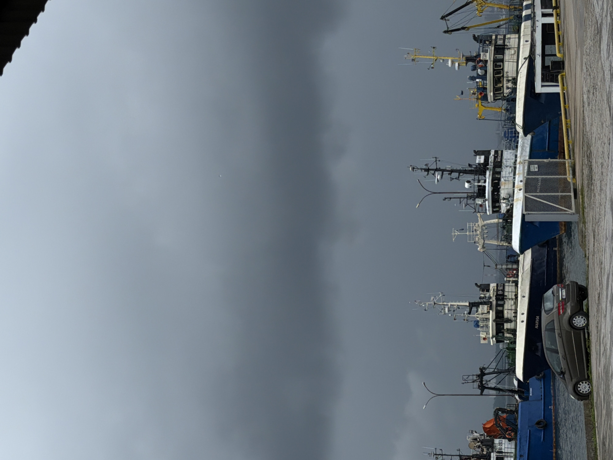 In the city of Viimsi, Estonia, a fleet of fishing vessels is docked at the harbor under a heavily overcast, grey sky, suggesting imminent rain or a storm. In the foreground, a dark grey Mercedes-Benz B-Class hatchback is parked on a paved area next to the water. The car is positioned at a slight angle towards the water, with its front facing left and its rear to the right. The vehicle's license plate is partially visible and appears to be of Estonian format.
Behind the car, several fishing boats are moored. The closest vessel on the right is a white and blue fishing trawler with the name "NARVIA" visible on its hull, and the registration number "B-5222" near its superstructure. It has a prominent mast and various deck equipment, including railings and what appears to be a small cabin. To the left of the "NARVIA," another similar fishing vessel, predominantly blue, is docked. This boat has a large orange object on its deck, possibly a rescue capsule or equipment. Further to the left, more fishing boats are partially visible. The harbor infrastructure includes docks, metal railings, and lighting poles. The overall scene conveys a sense of industrial maritime activity in a potentially challenging weather environment.