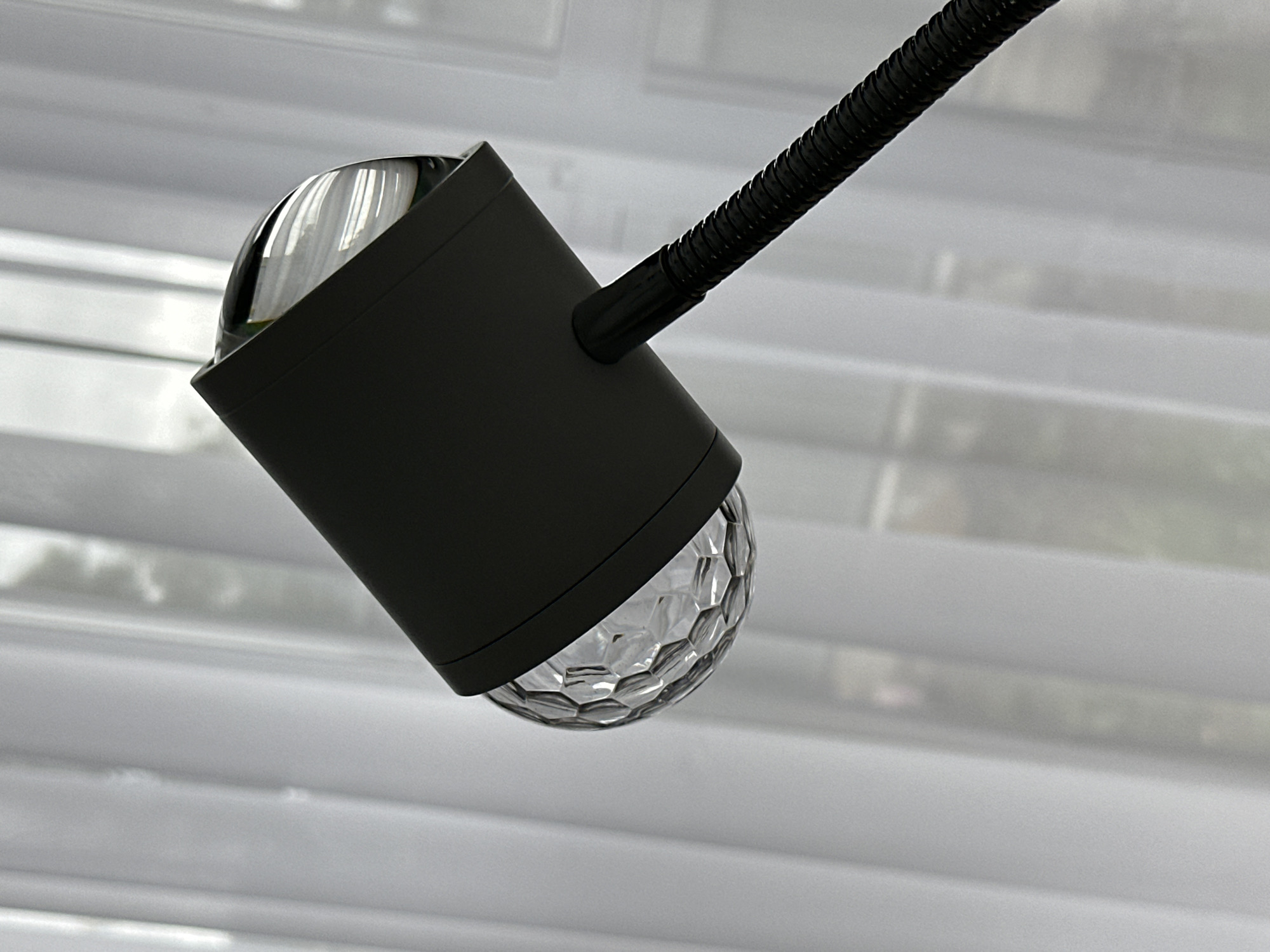 A black desk lamp with a clear multifaceted bulb is positioned in front of white Venetian blinds. The lamp has a cylindrical shade, a gooseneck arm, and a partially visible clear dome-shaped lens. The location appears to be indoors.