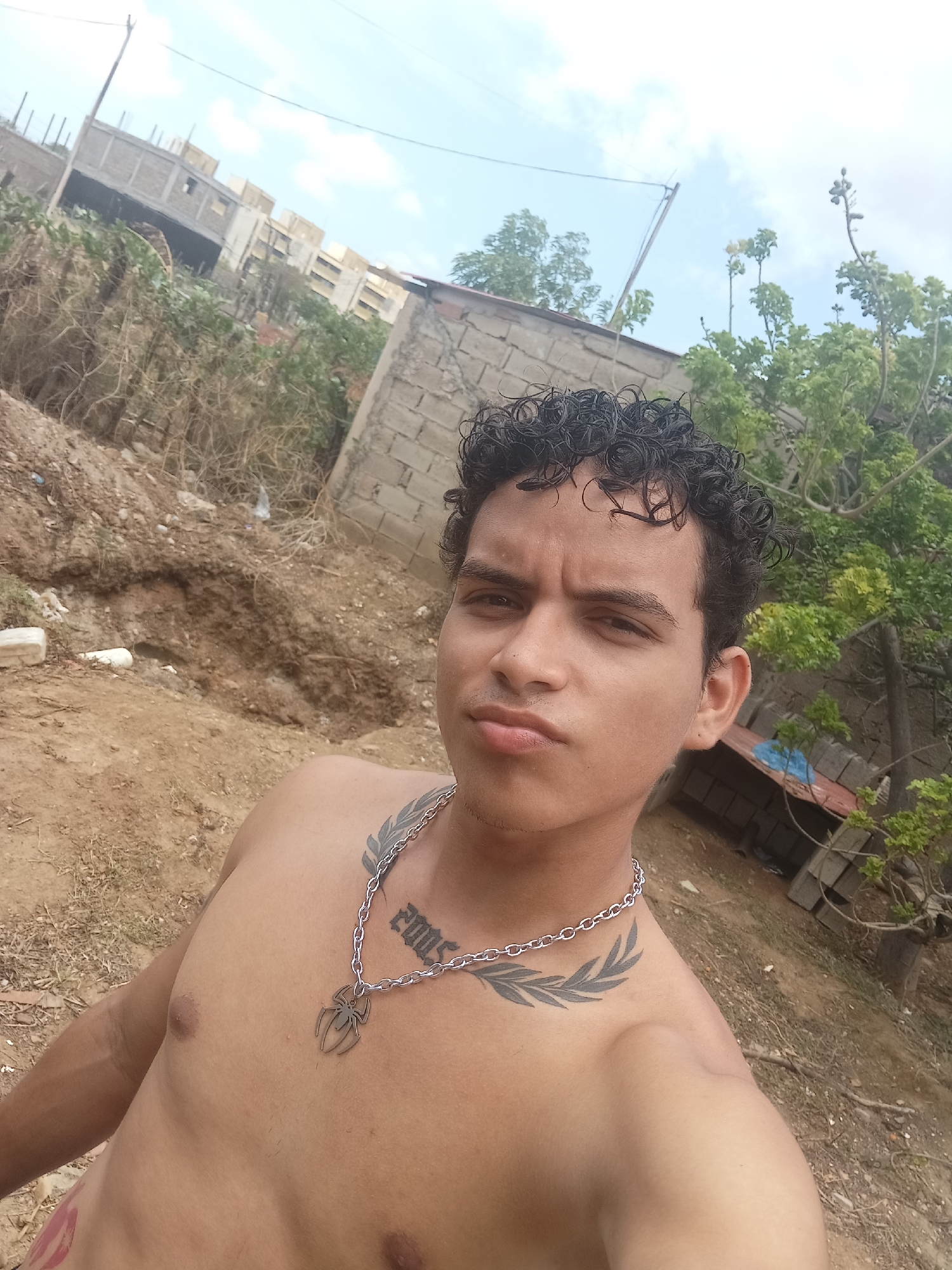 A young man, appearing to be in his late teens or early twenties, with dark, curly hair, poses for a selfie outdoors. He is shirtless, revealing a tattoo of a spider on a chain necklace around his neck and a leafy design tattooed on his left shoulder and upper chest. His facial expression is serious, with pursed lips. The background suggests an urban or semi-urban environment in Altagracia, Venezuela, featuring unfinished construction, rubble, dry vegetation, and a bright, overcast sky. The ground is a mixture of dirt and dry grass. To the right, a rudimentary wooden structure with a blue tarp is visible. The image captures a candid moment in what appears to be an undeveloped or construction-heavy area.