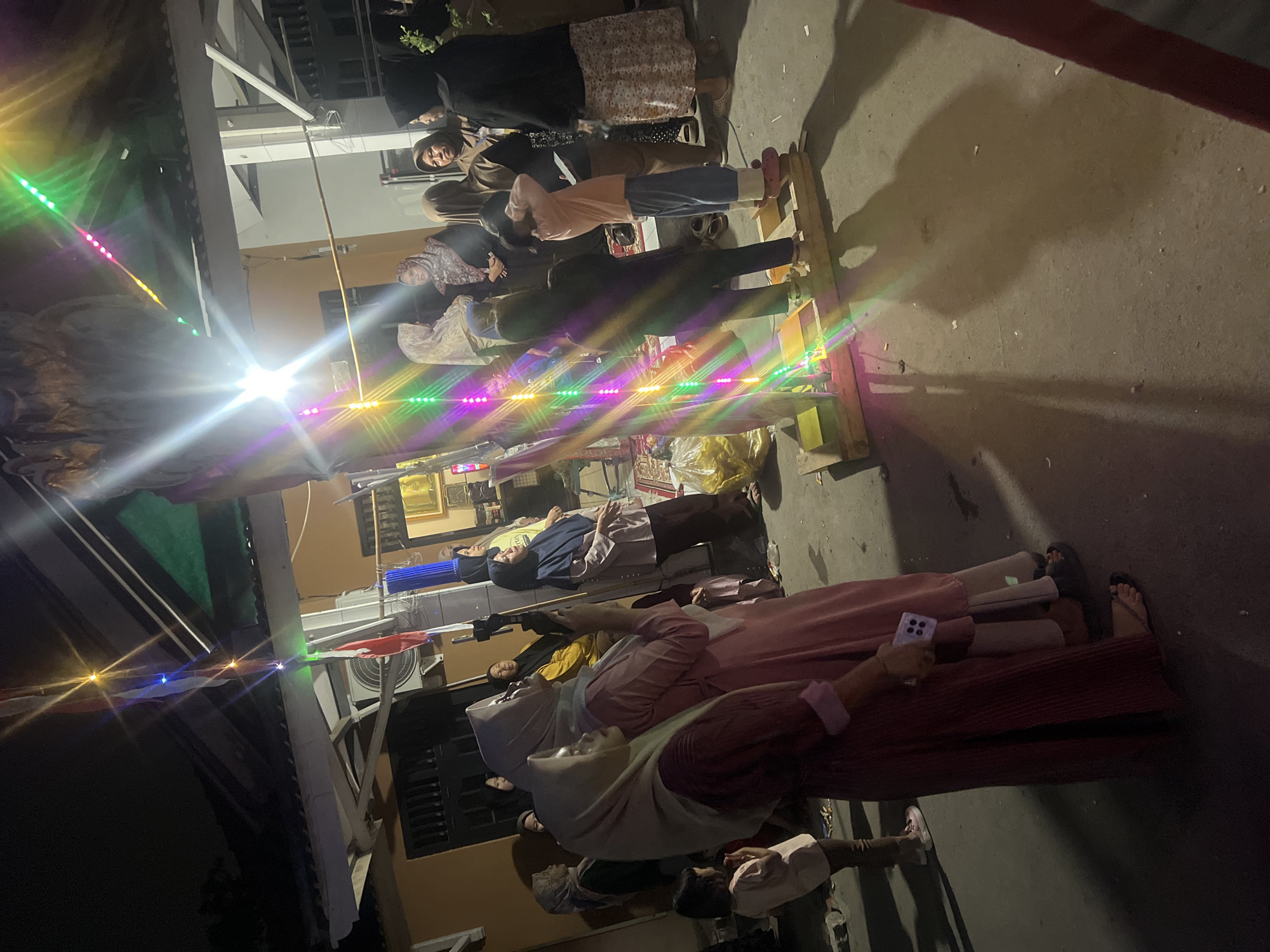 The image depicts a night scene in Klapanunggal, Indonesia, likely during a local celebration or gathering. A group of people, primarily women and children, are gathered around a temporary wooden structure adorned with colorful LED lights. Some individuals are holding up mobile phones, possibly recording or taking photos. The environment appears to be an outdoor area, possibly a yard or street, with a building visible in the background. The lighting is dim, with the LED lights and a bright light source creating lens flares and casting shadows. The overall atmosphere suggests a community event taking place after dark.