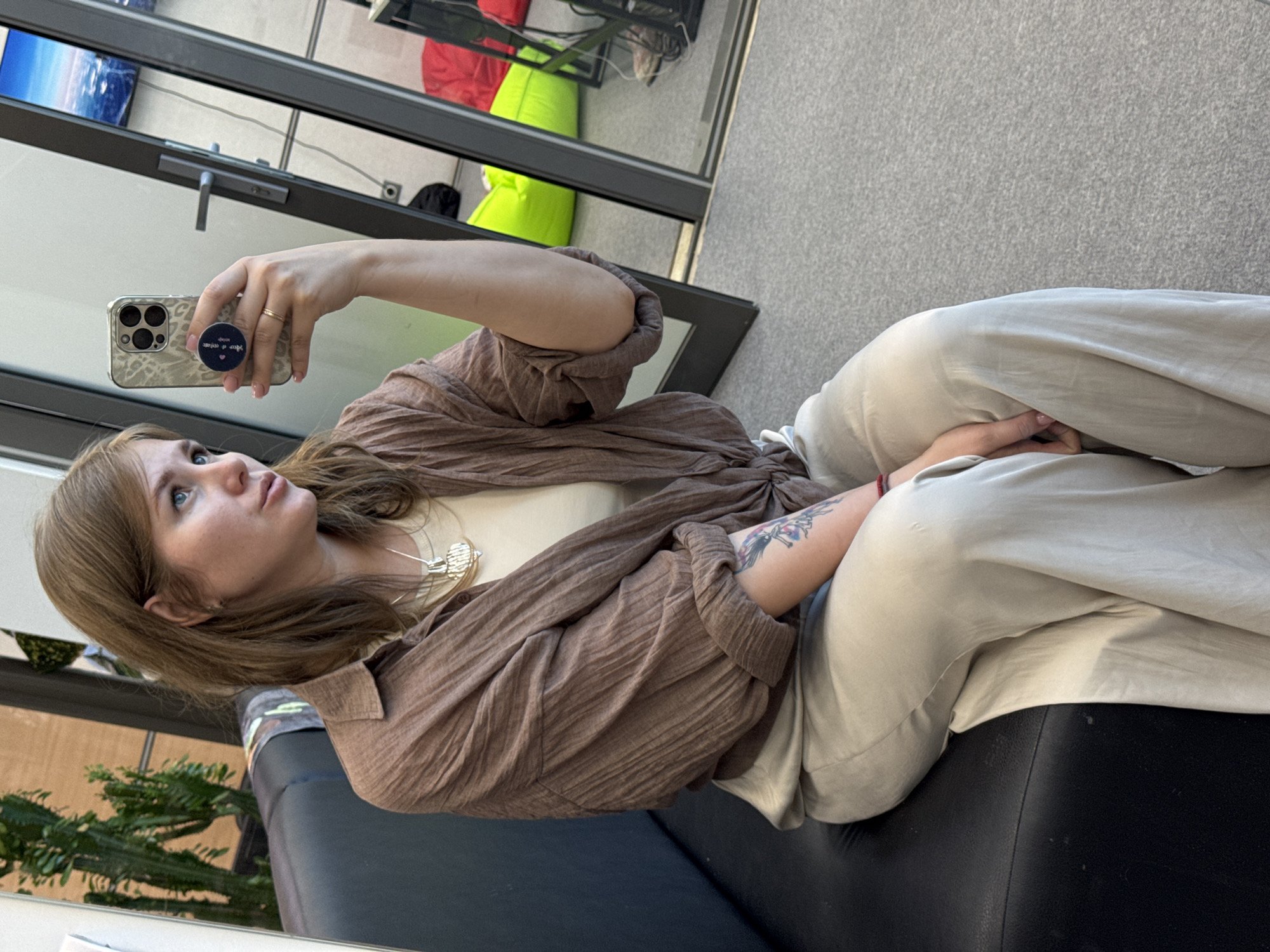 A woman sits on a black leather sofa. She is fair-skinned, with light brown hair, and blue eyes. She holds a cell phone in her left hand, presumably taking a selfie. She is wearing light-colored pants and a brown, textured long-sleeved shirt over a light-colored top. A necklace is visible. Her left arm has a tattoo.
Behind her, a potted plant is visible. Through a window in the background, there are glimpses of green and red items, suggesting an interior setting. Based on the phone case with the inscription in Russian and the presence of a Yandex identifier, it is likely this photo was taken in Russia. As such, it is possible that this picture was taken in the city of Perm, Russia.
