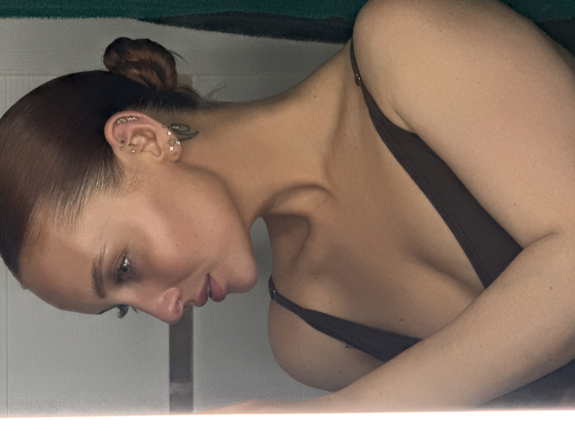 A woman with her hair pulled into a bun is depicted in a close-up, slightly angled shot. She is wearing a dark-colored, strapped garment, potentially a swimsuit or top. Her head is turned downwards, drawing attention to her neck and collarbone. Her left ear is adorned with multiple piercings. The background appears to be a sparsely decorated interior space with a white wall and a visible dark green surface above. The image was taken indoors, but there is no definitive way to tell whether the image was taken in Chaikovsky, Russia.