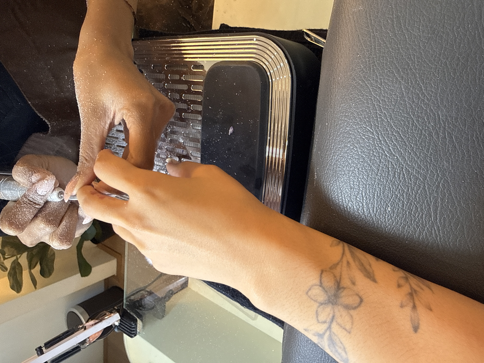 The image shows a close-up of two hands during a nail care procedure. The hand on the right is being worked on by a nail technician, who is using an electric nail file. Dust from the filing process is visible on the hands and the table. The hand on the left belongs to the person receiving the nail treatment and has a floral tattoo on the forearm. The scene appears to be taking place in a salon or spa setting, with a black leather chair in the foreground and a potted plant and a trash can visible in the blurred background. The environment is indoors, with artificial lighting. The location is specified as North Kuta, Indonesia.