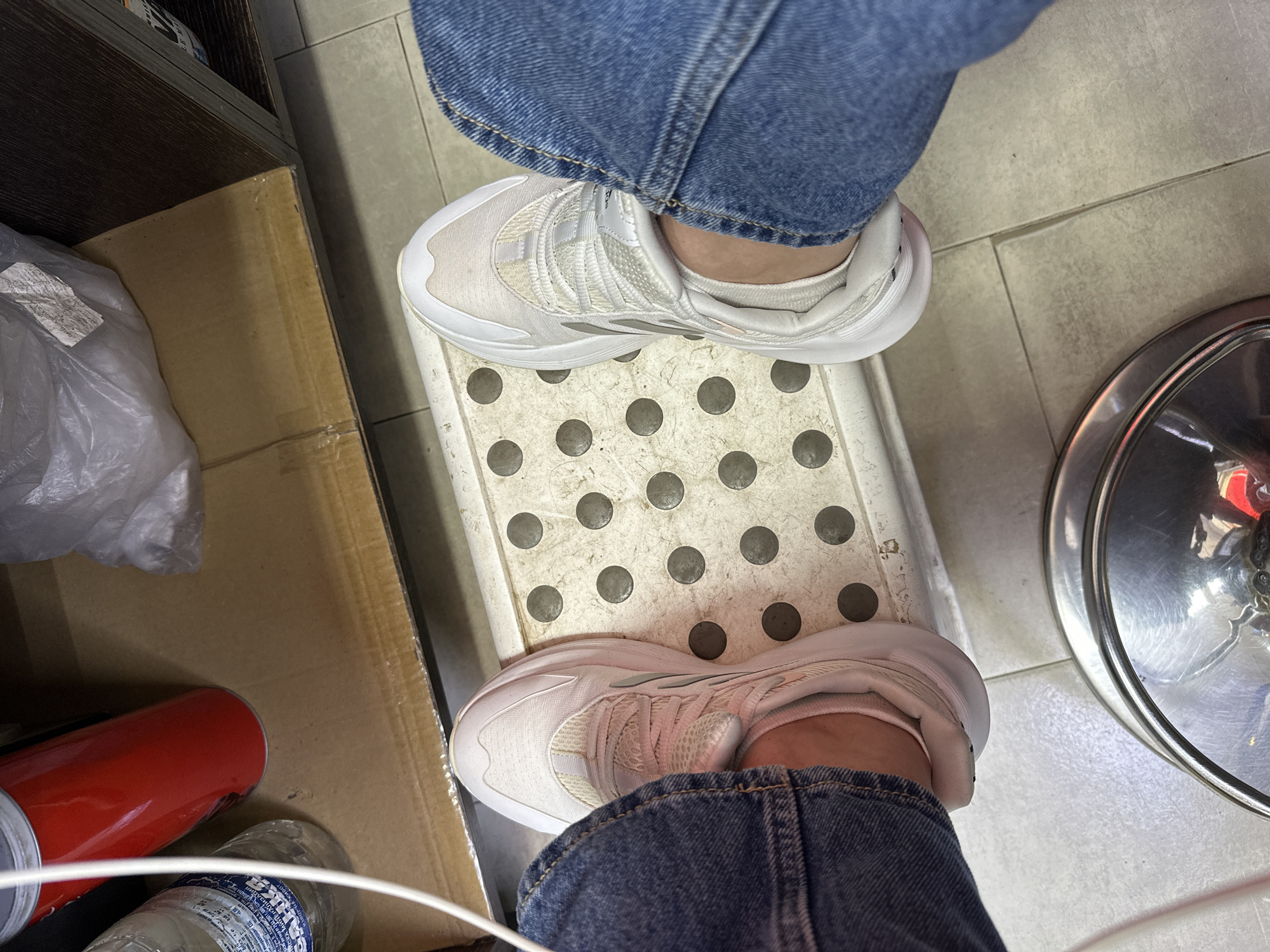 The image shows a person's feet wearing white athletic shoes and blue jeans. The person is standing on a white plastic step stool with circular indentations. In the background, there is a cardboard box, a red cylindrical object, and a white plastic bag. The floor appears to be tiled. The scene suggests an indoor setting, possibly a storage area or a workshop, and is not identifiable as being in Sofia, Bulgaria.