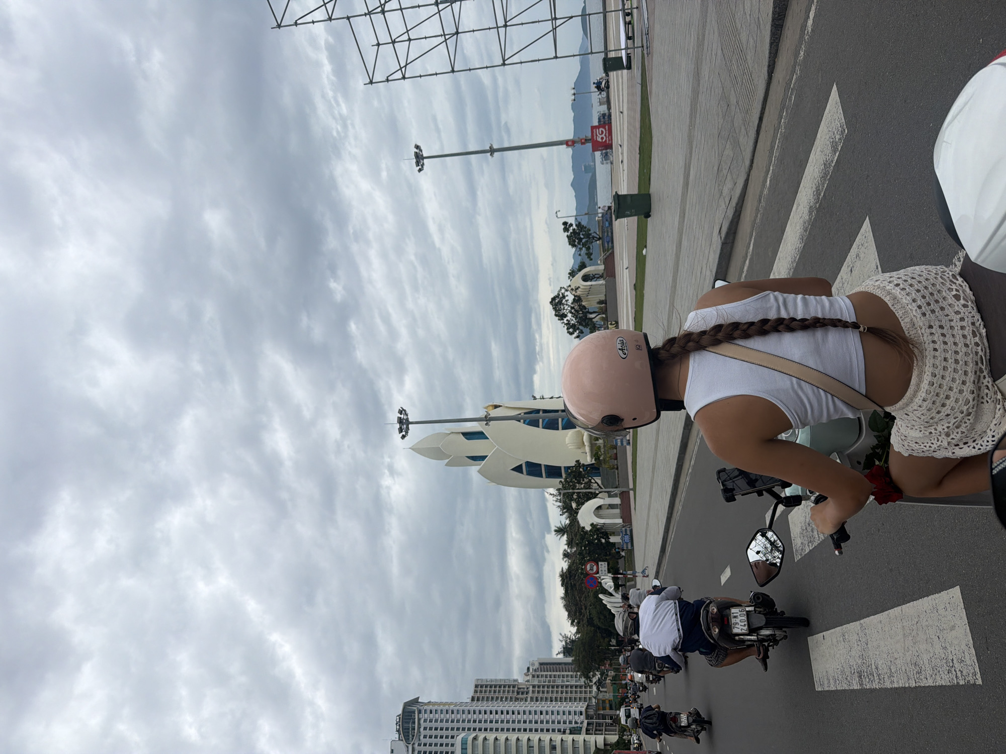 This is a photograph taken from the perspective of someone riding on the back of a motorcycle in Nha Trang, Vietnam. The rider in front is wearing a white tank top and a crocheted white skirt. She has a pink helmet on and a long braid of brown hair. She is holding the handlebars of the motorcycle. In front of them is a busy street with many other motorcycles and a few cars. The street is lined with tall buildings on the left and a waterfront promenade on the right. In the background, a distinctive lotus-shaped building with white petals and blue windows stands out. The sky is overcast with gray clouds. There is a large metal scaffolding structure on the far right side of the image. The road has white crosswalk markings.