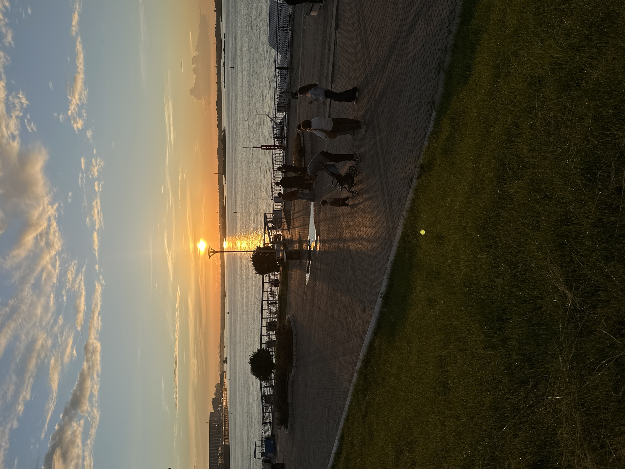 This image depicts a sunset scene on a waterfront promenade in an unidentified city. The sun, a brilliant orb, is low on the horizon, casting a warm, golden glow across the sky and water. Scattered clouds, tinged with orange and pink, drift across the blue expanse.
In the foreground, a paved promenade, made of interlocking bricks, stretches out. To the left, a sloped grassy area borders the promenade. A dark metal railing runs along the edge of the promenade, separating it from the water. Beyond the railing, a wide river or bay reflects the sunset. On the opposite bank, a cityscape with industrial buildings and a bridge is visible.
Several people are present on the promenade. In the mid-ground, a group of individuals are gathered, including a couple with a baby in a stroller, and another couple. Further back, closer to the railing, are more silhouettes of people. A lamppost with a bright light is visible, its reflection shimmering on the wet pavement.
The overall atmosphere is peaceful and serene, with the warm light of the setting sun creating a picturesque scene. The image captures a moment of everyday life by the water, with people enjoying the view and each other's company.