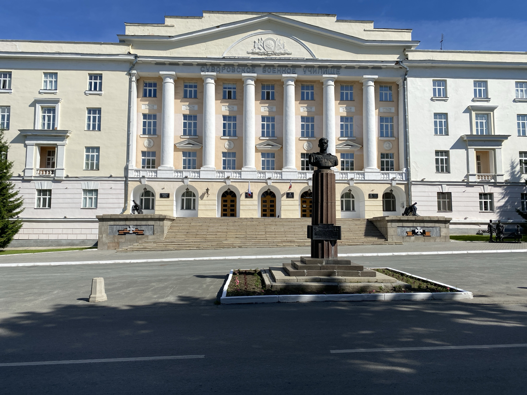 The image shows a frontal view of the Yekaterinburg Suvorov Military School in Yekaterinburg, Russia. The school is a large, imposing, pale yellow building with classical architectural elements, including a colonnade of large white pillars across the facade. Above the pillars, the inscription "СУВОРОВСКОЕ ВОЕННОЕ УЧИЛИЩЕ" (Suvorov Military School) is visible in white lettering. A large pediment with a crest adorns the center of the building's top.
In front of the building, a prominent monument featuring a bust of a military figure on a pedestal stands centrally. The pedestal is adorned with a plaque. Steps lead up to the main entrance of the building. To the left and right of the monument base, there are decorative stone structures, one of which appears to have a cannon and another with two figures.
The foreground shows a paved asphalt area, likely a parade ground or street, with white lane markings. A small, light-colored concrete bollard is situated in the middle of the asphalt. The scene is illuminated by bright sunlight, casting distinct shadows on the ground. A lush green coniferous tree is visible to the far left of the building. The sky is a clear, bright blue.