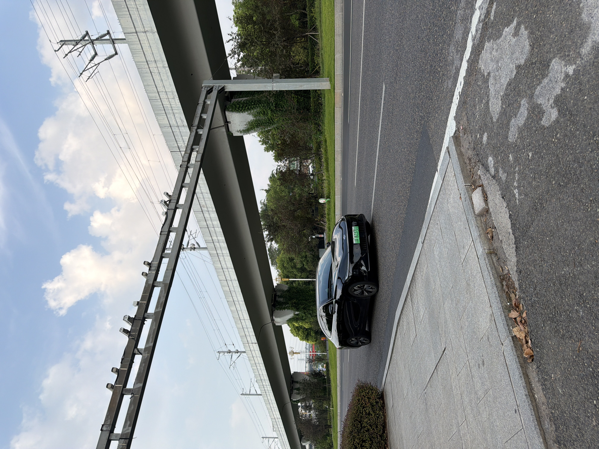 A black electric car is driving on an asphalt road under a large elevated structure, likely an overpass or elevated railway. The car has a green license plate with the characters "A BZ3701". The elevated structure is supported by concrete pillars that are partially covered in ivy. Overhead power lines are visible, suggesting the presence of a railway or other electrical infrastructure. The sky is partly cloudy with scattered white clouds against a light blue background. To the left of the road is a sidewalk with large granite tiles and a low, manicured hedge. Trees and other greenery line the area beyond the road. The scene appears to be in an urban or suburban environment. Based on the license plate format and the visible infrastructure, this scene is likely located in Jiangjin District, Chongqing, China.