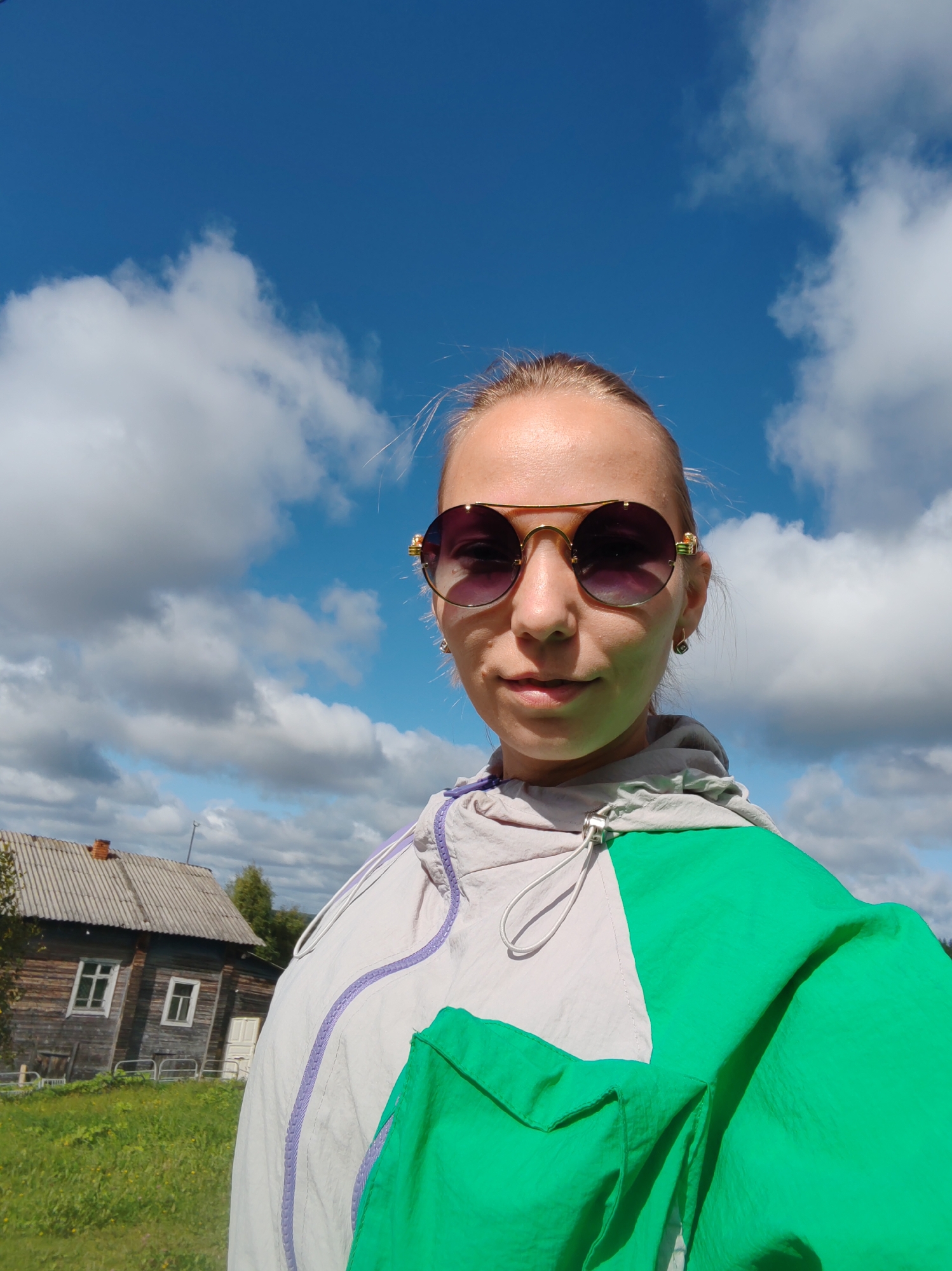 A young woman with blonde hair pulled back and wearing sunglasses, a light gray and green jacket, and an earring, takes a selfie outdoors. She is positioned in the foreground, with a wooden house and a field of grass behind her. The sky above is blue with white clouds. The scene appears to be in a rural area, potentially the city of Chuchlam, Russia, given the style of the house and the natural surroundings.